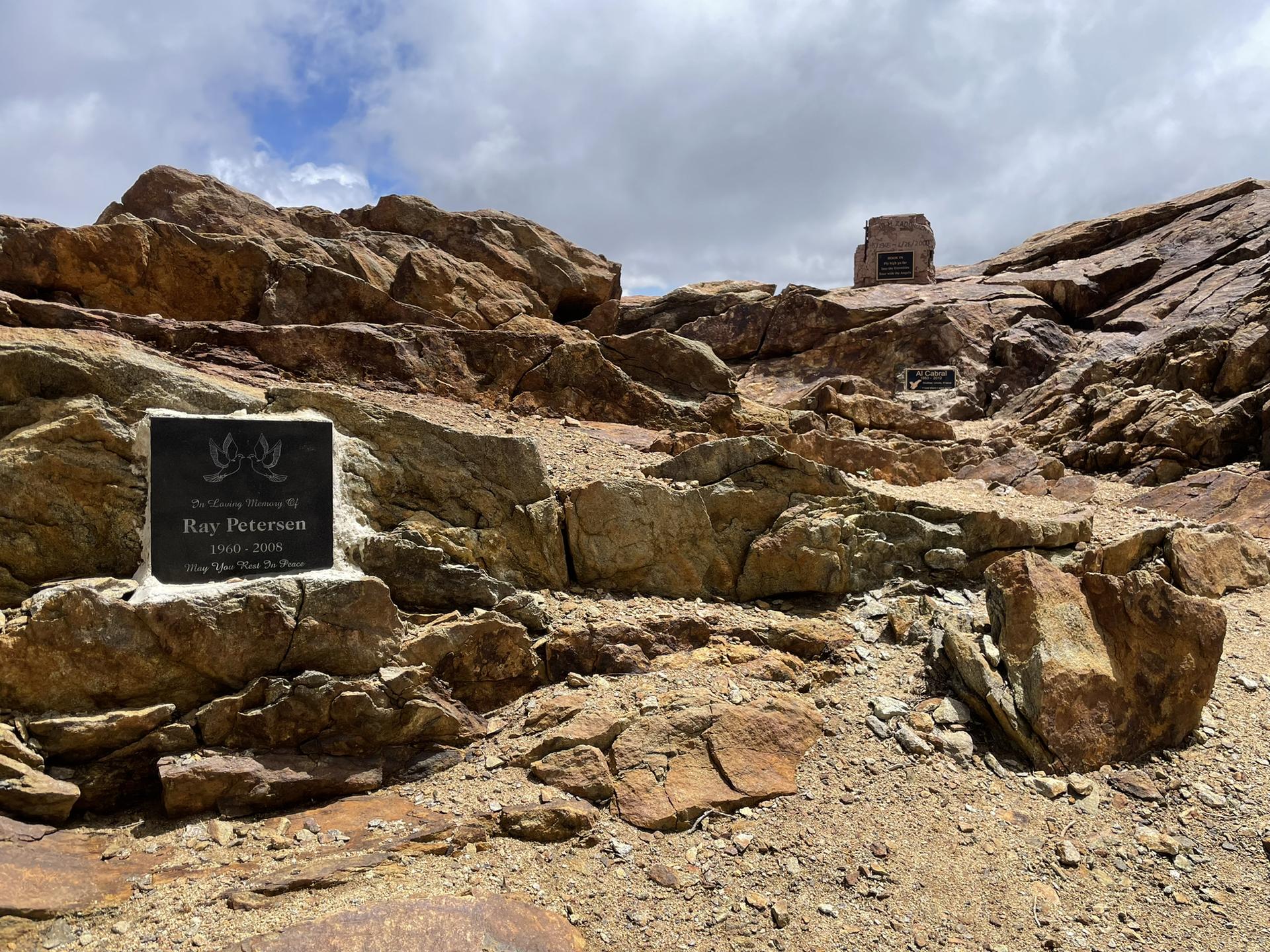 Memorial plaques on rocks in a rugged, rocky mountain landscape under a cloudy sky.