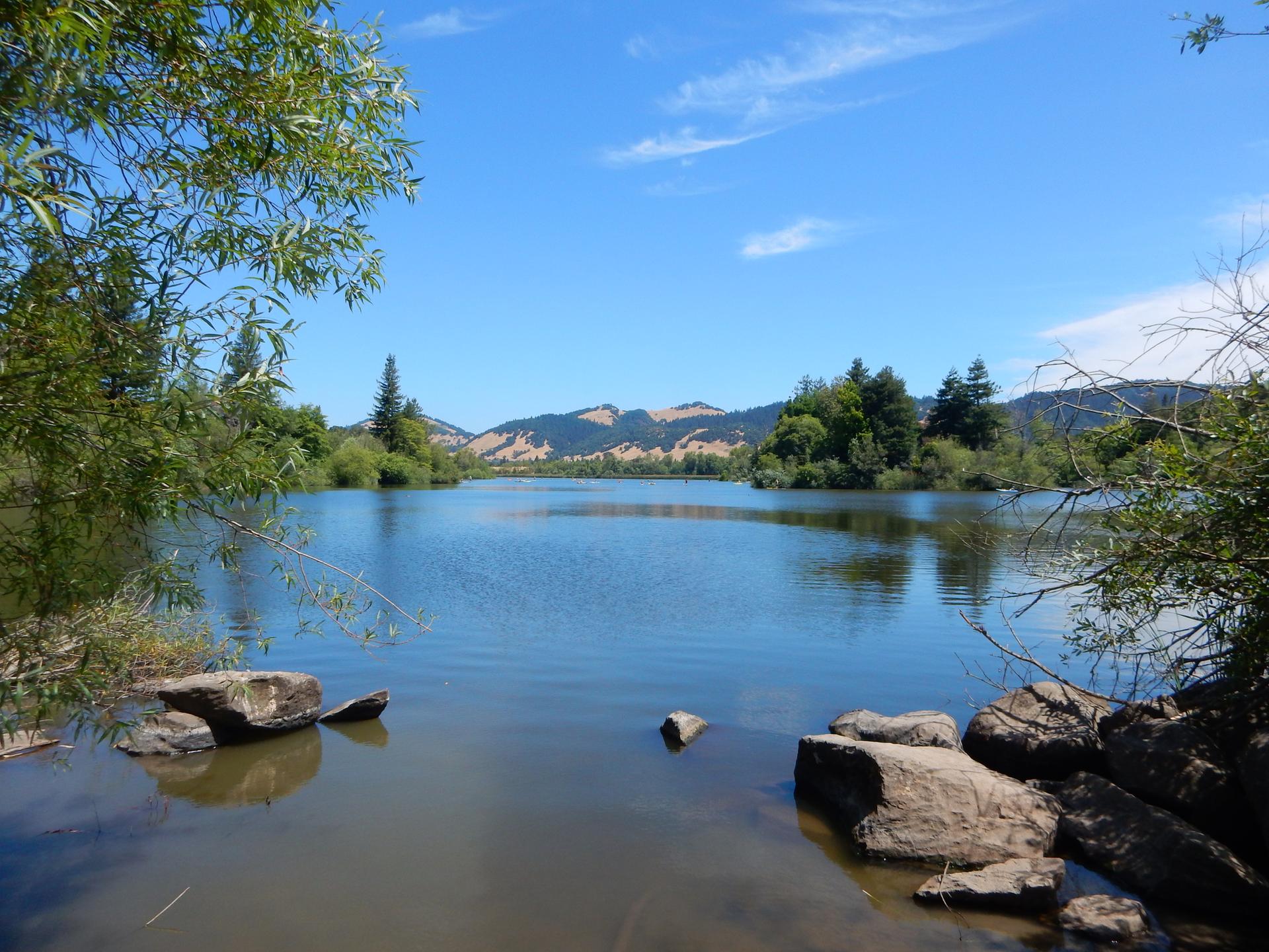 Calm lake with rocks in the foreground, distant hills under a blue sky.