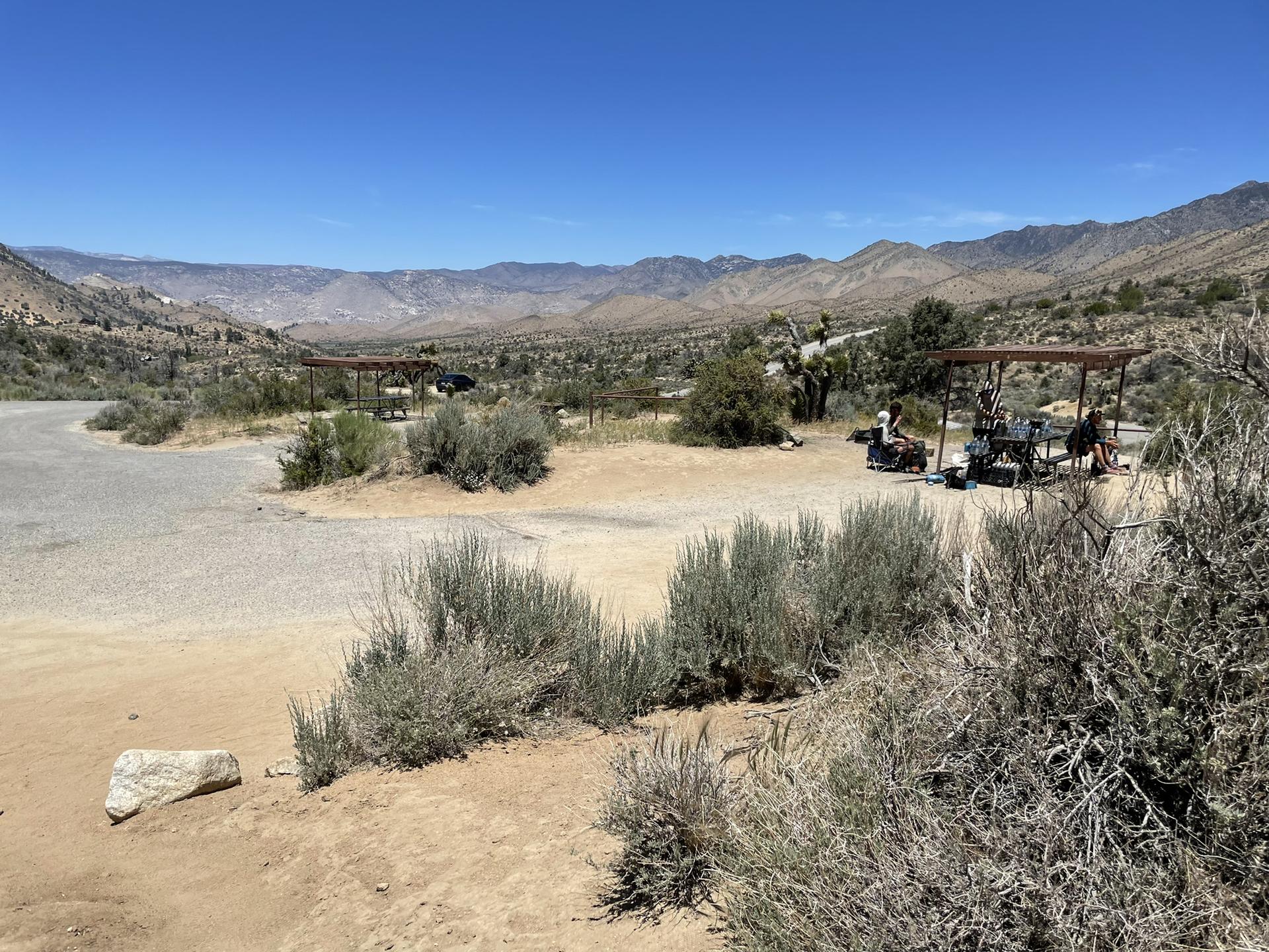 A wide, sunny desert landscape with distant mountains and two shaded picnic areas.