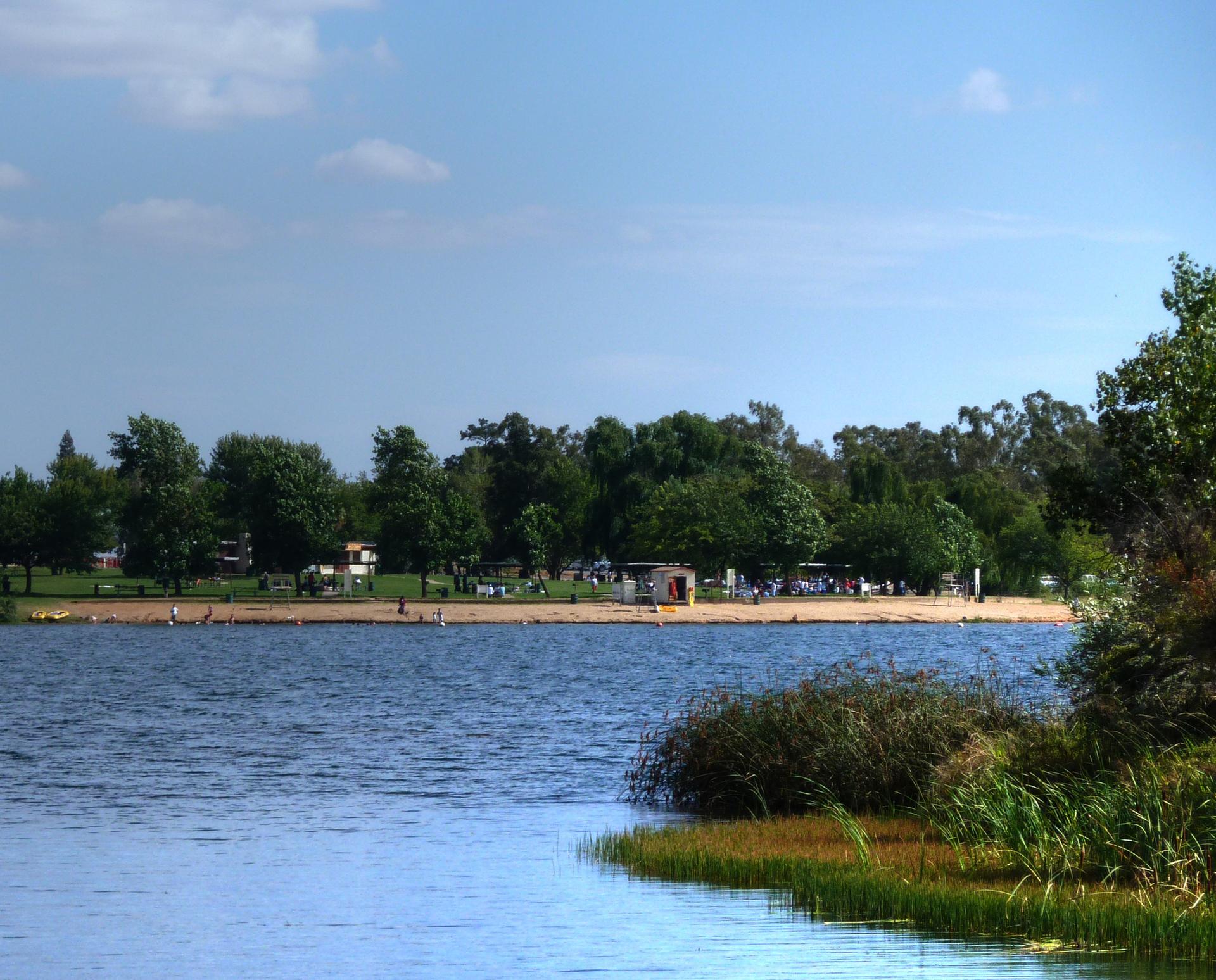 A sunny lake with a sandy beach, distant trees, and people enjoying the water.
