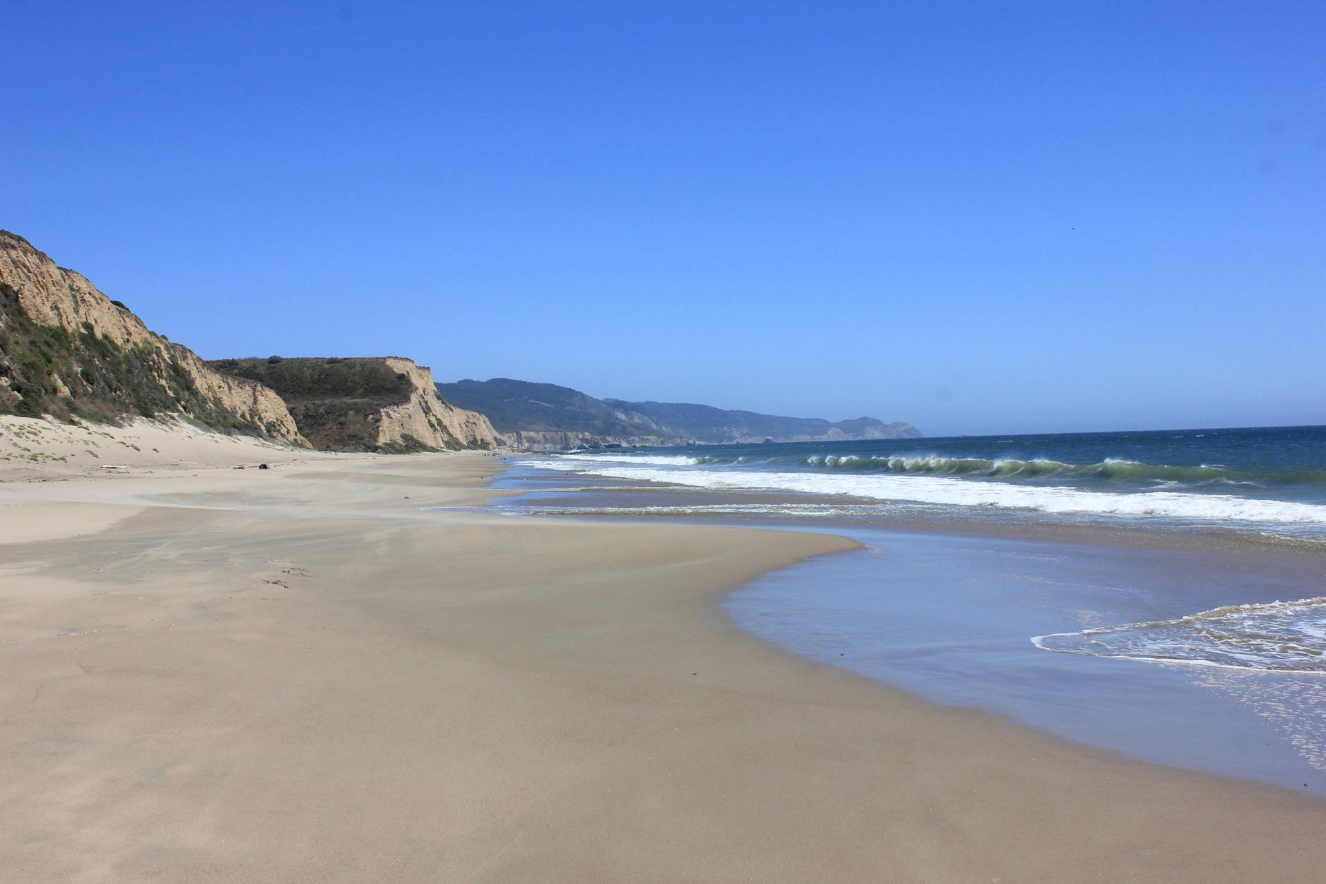 Sunny beach with ocean waves, sandy shore, and tall cliffs under blue sky.