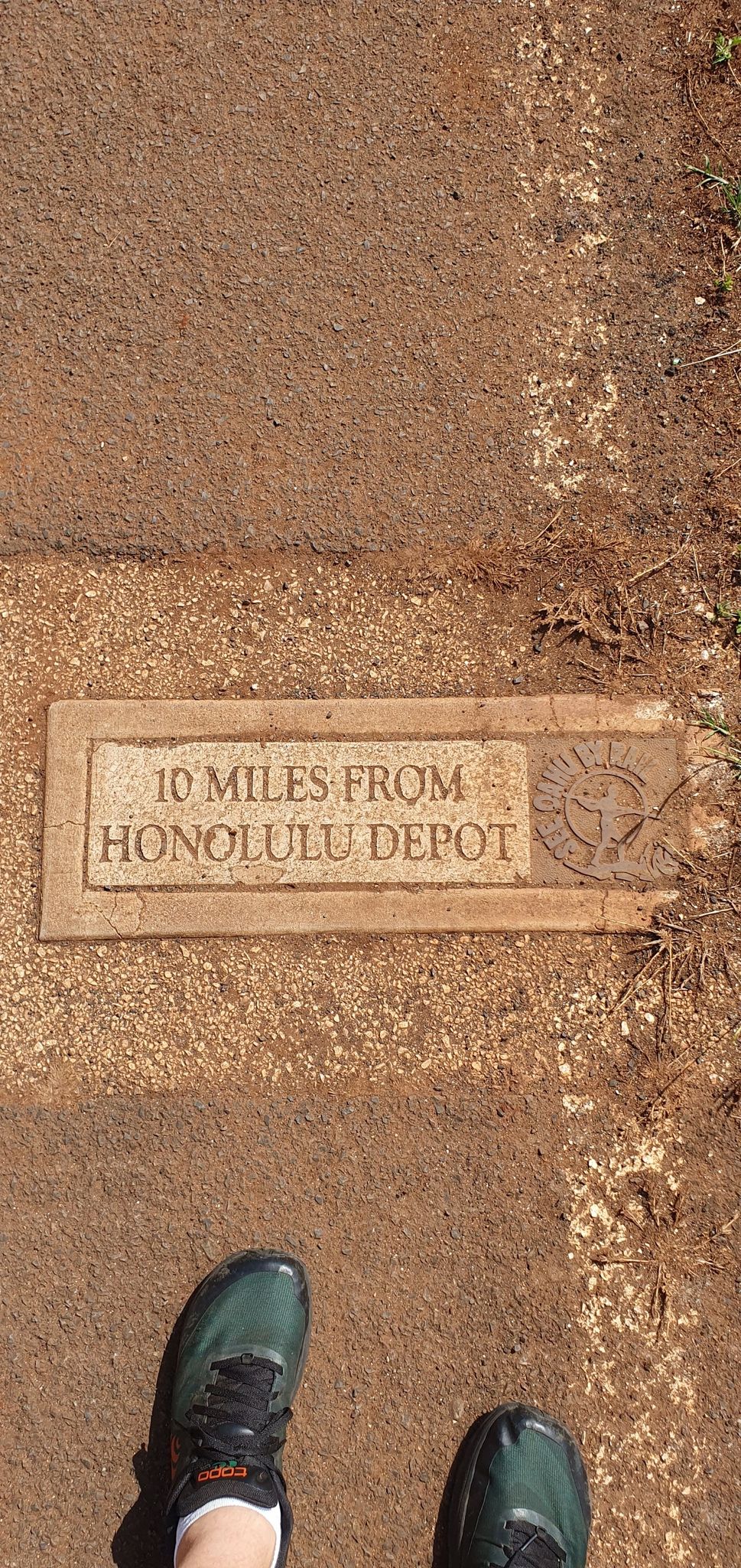 Feet in athletic shoes stand on a dirt path next to a stone marker. - Bike loop from Kalauao / Pearlridge