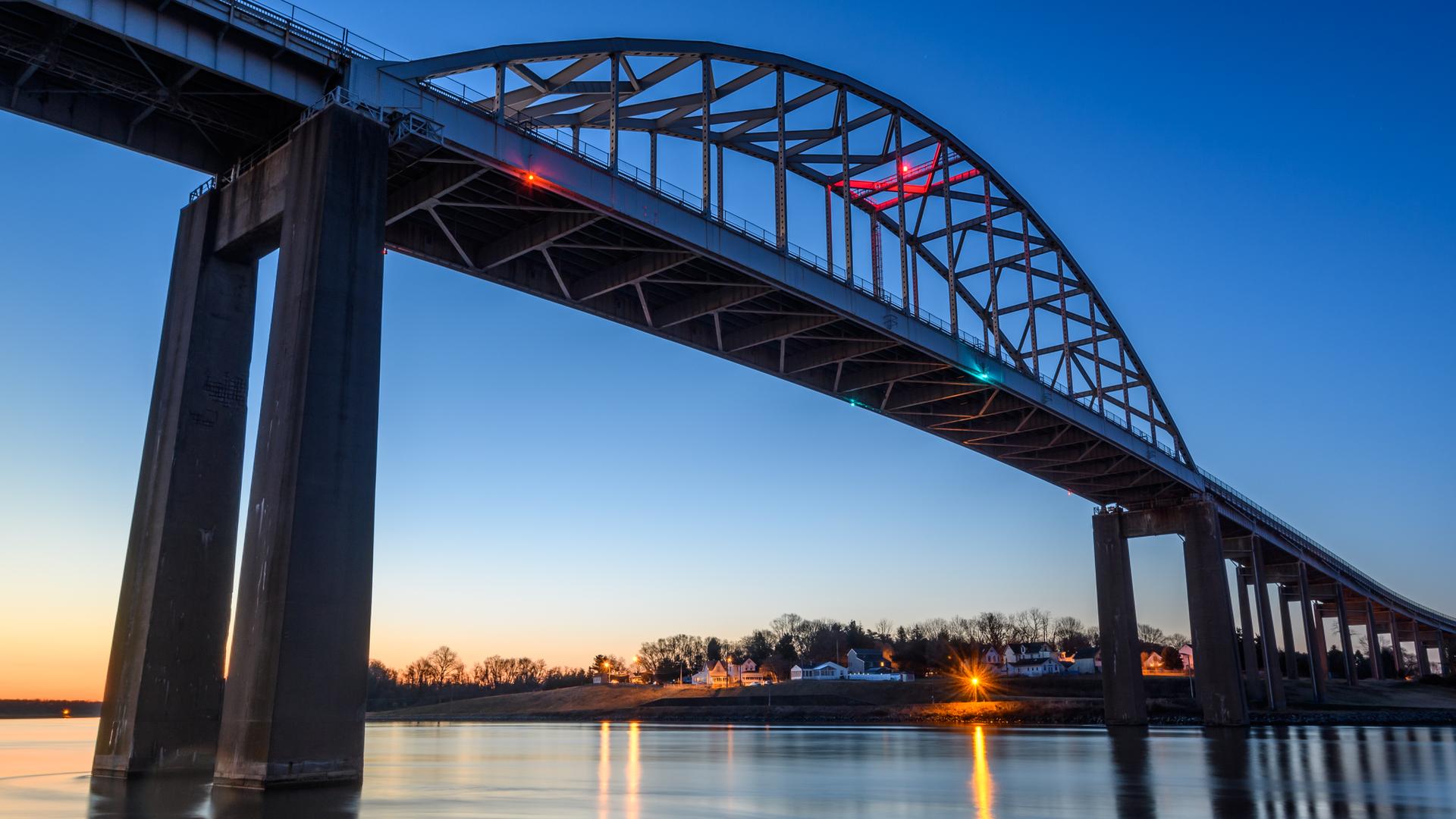 Arched bridge illuminated at dusk over water with distant shore lights.