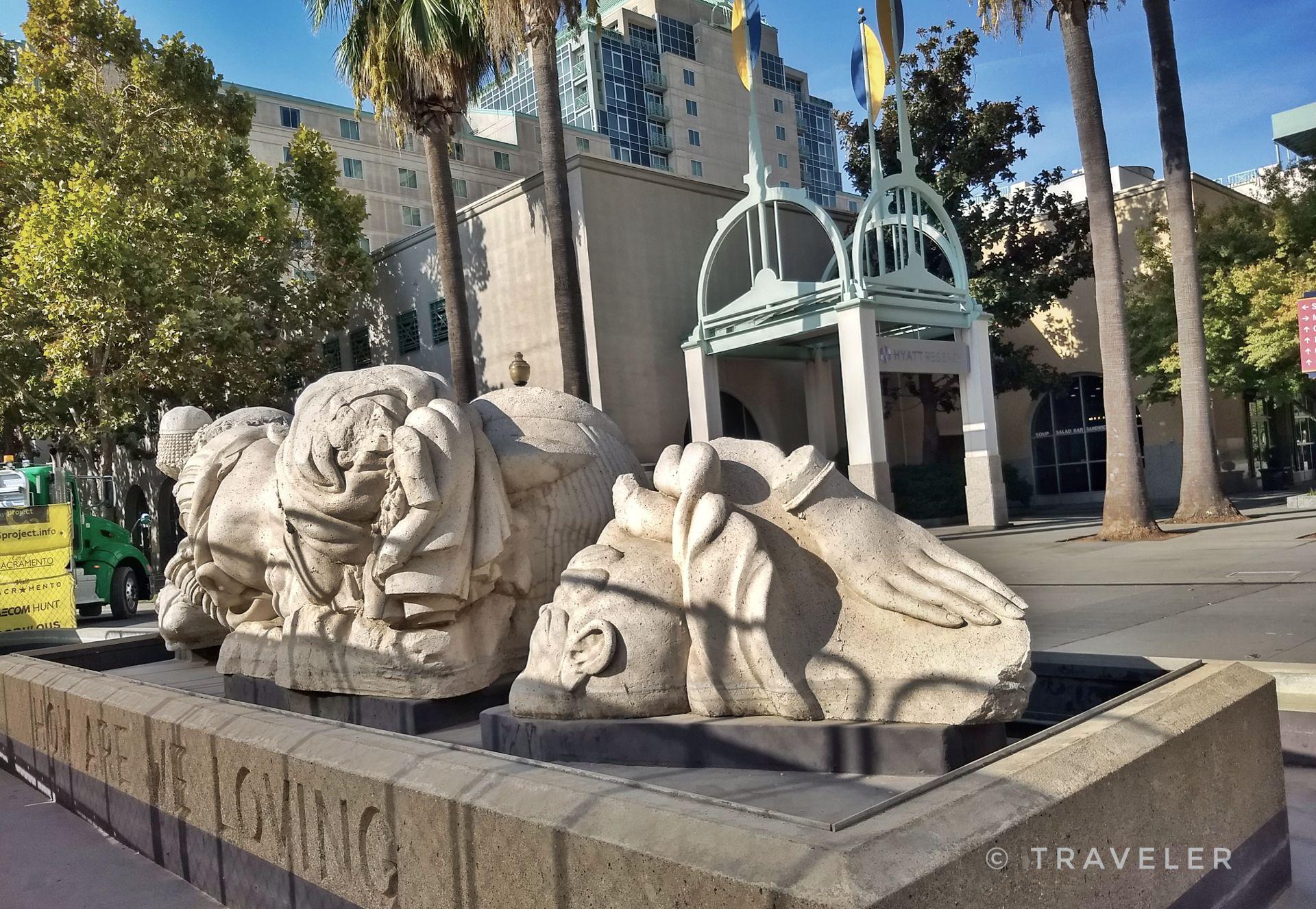 Large stone sculptures in a fountain, with palm trees and buildings under bright sun.