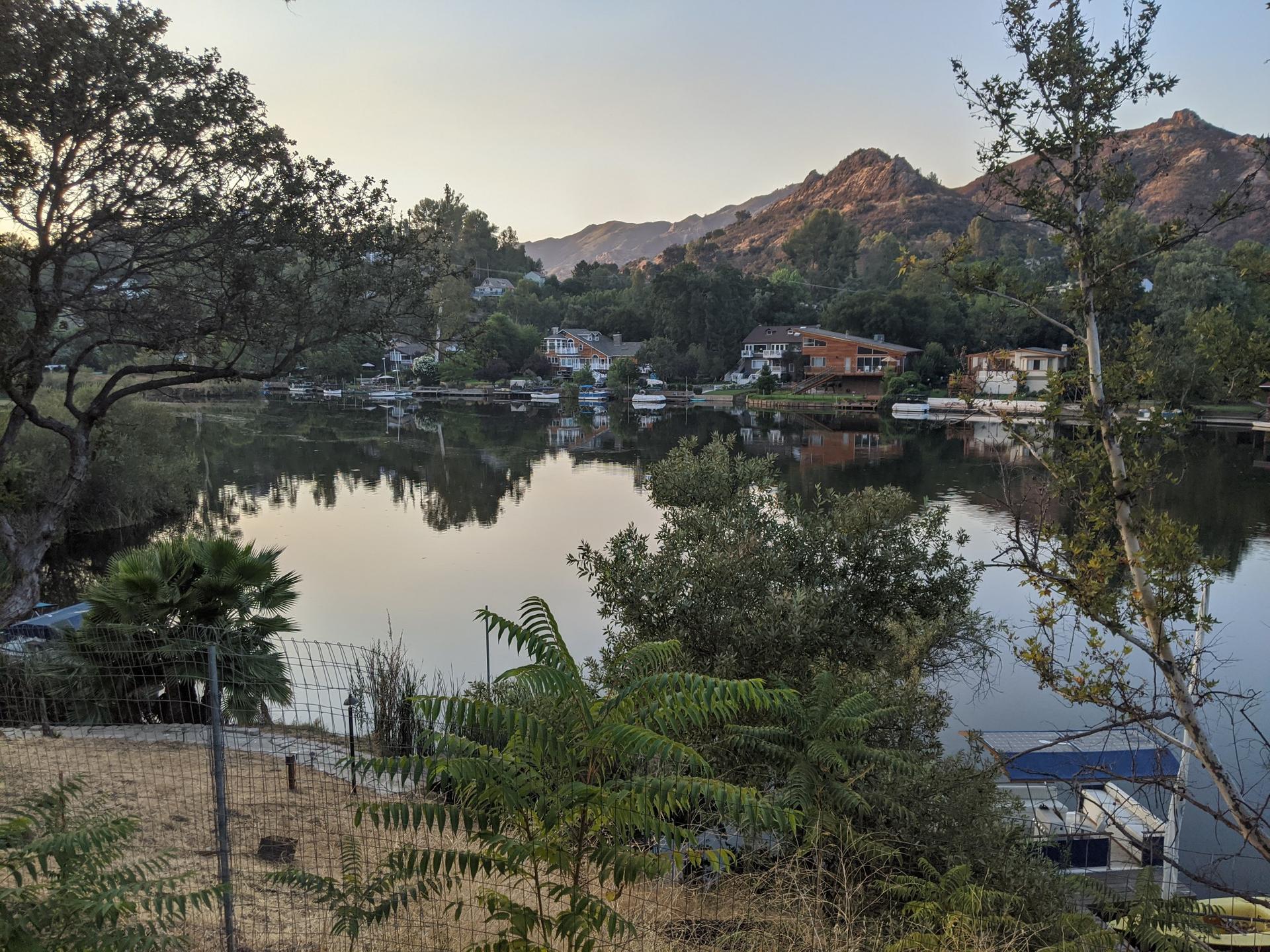 Calm lake reflecting houses and trees, with distant mountains at dusk.