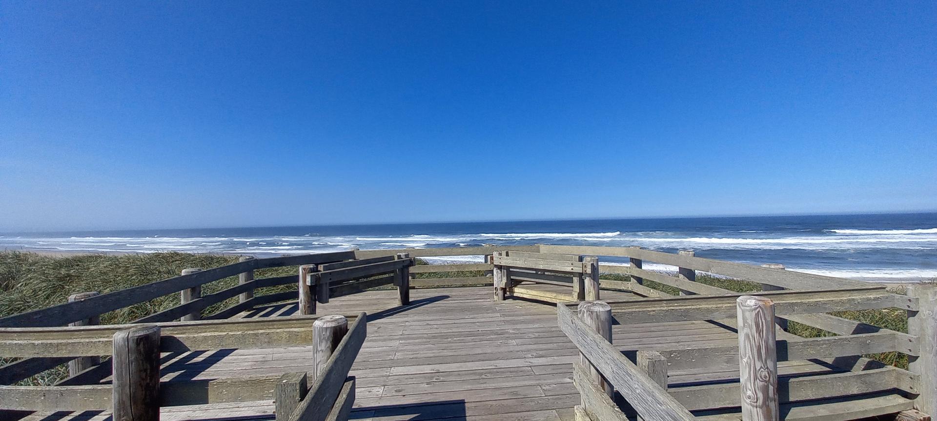 Wooden boardwalk overlooking a sunny beach and ocean under a clear blue sky.