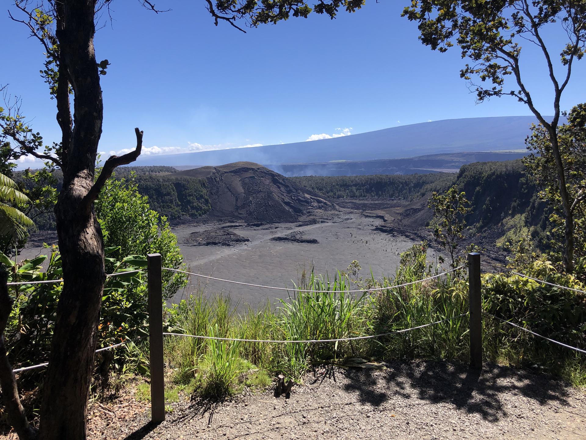 Bright day over a volcanic crater, framed by green trees and a rope fence.