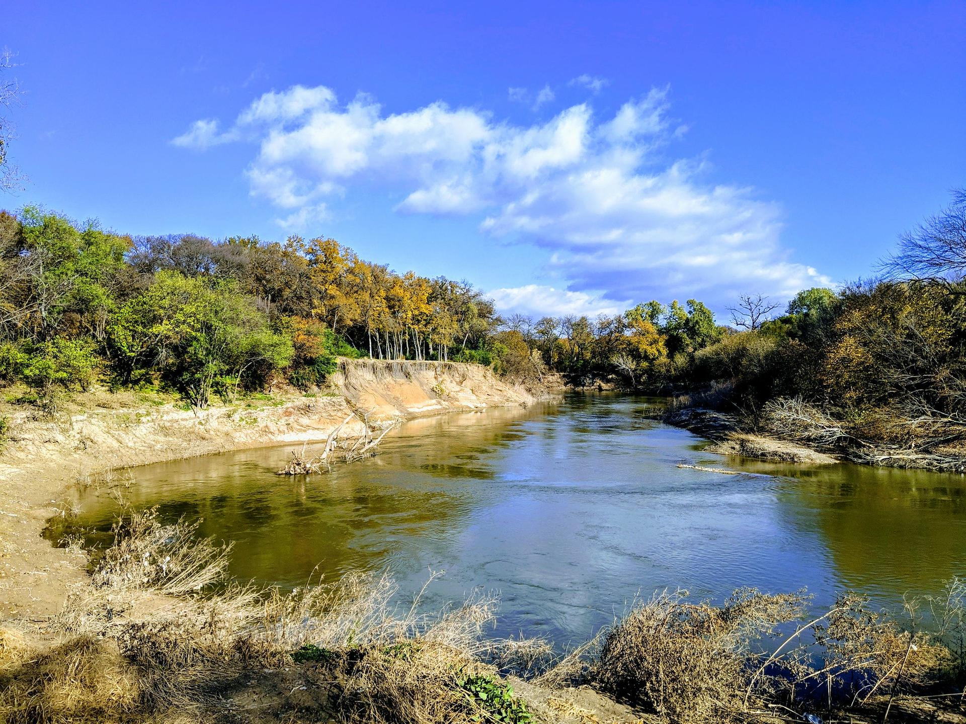 Wide river reflecting blue sky, surrounded by autumn trees and sandy banks.
