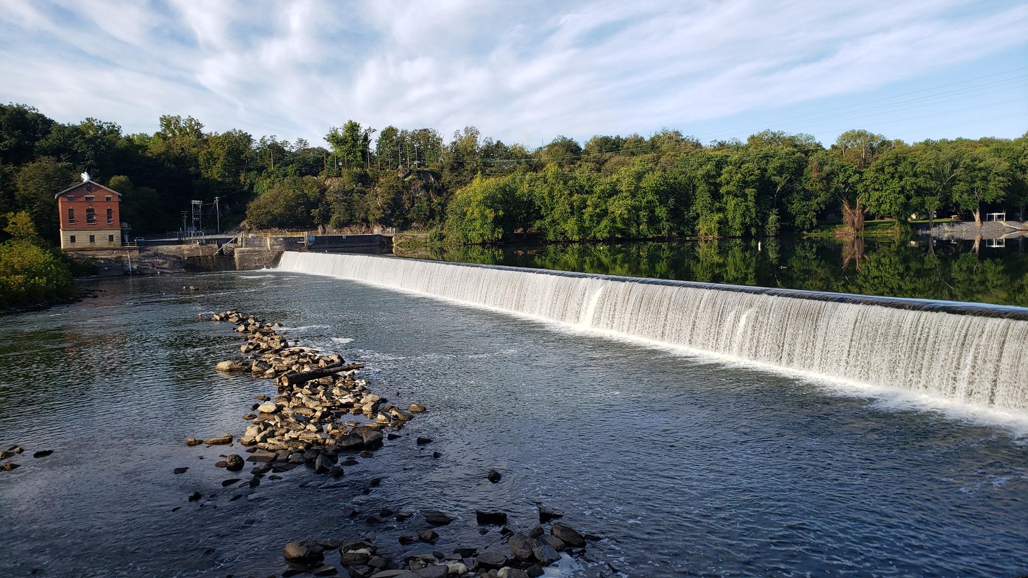 A wide dam creates a waterfall across a river, with a building nearby.