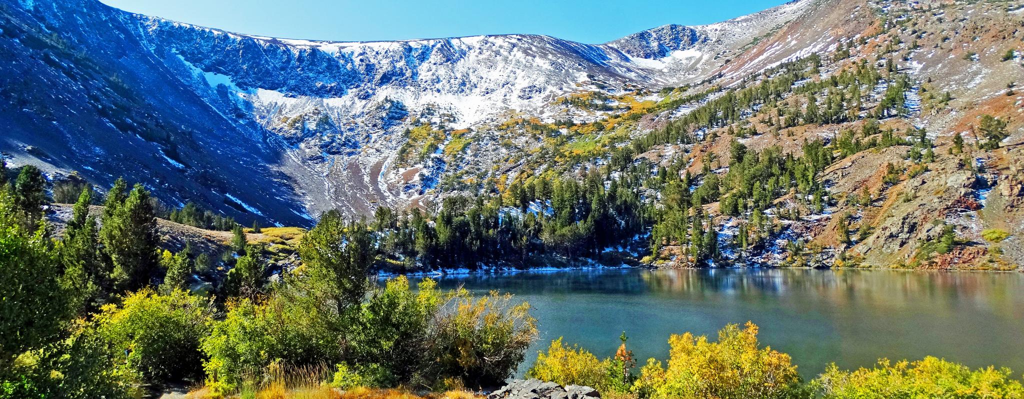 Mountain lake with snow-dusted peaks and colorful autumn trees under a clear sky.
