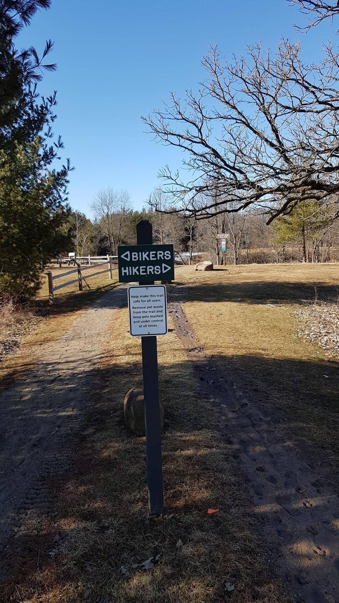 Trail sign for bikers and hikers on a dirt path on a sunny day.