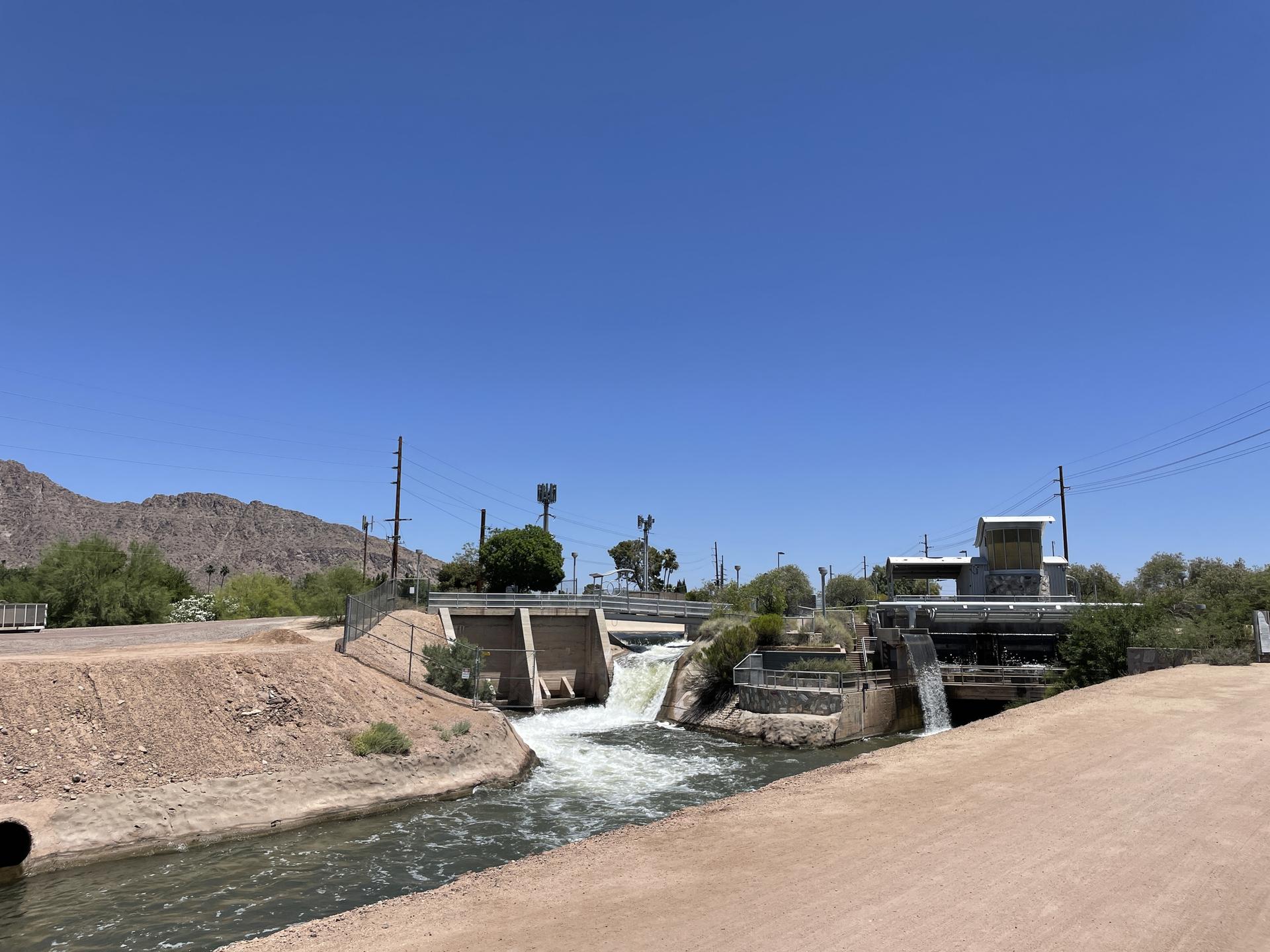 Water flows through a concrete dam structure with mountains in the background.