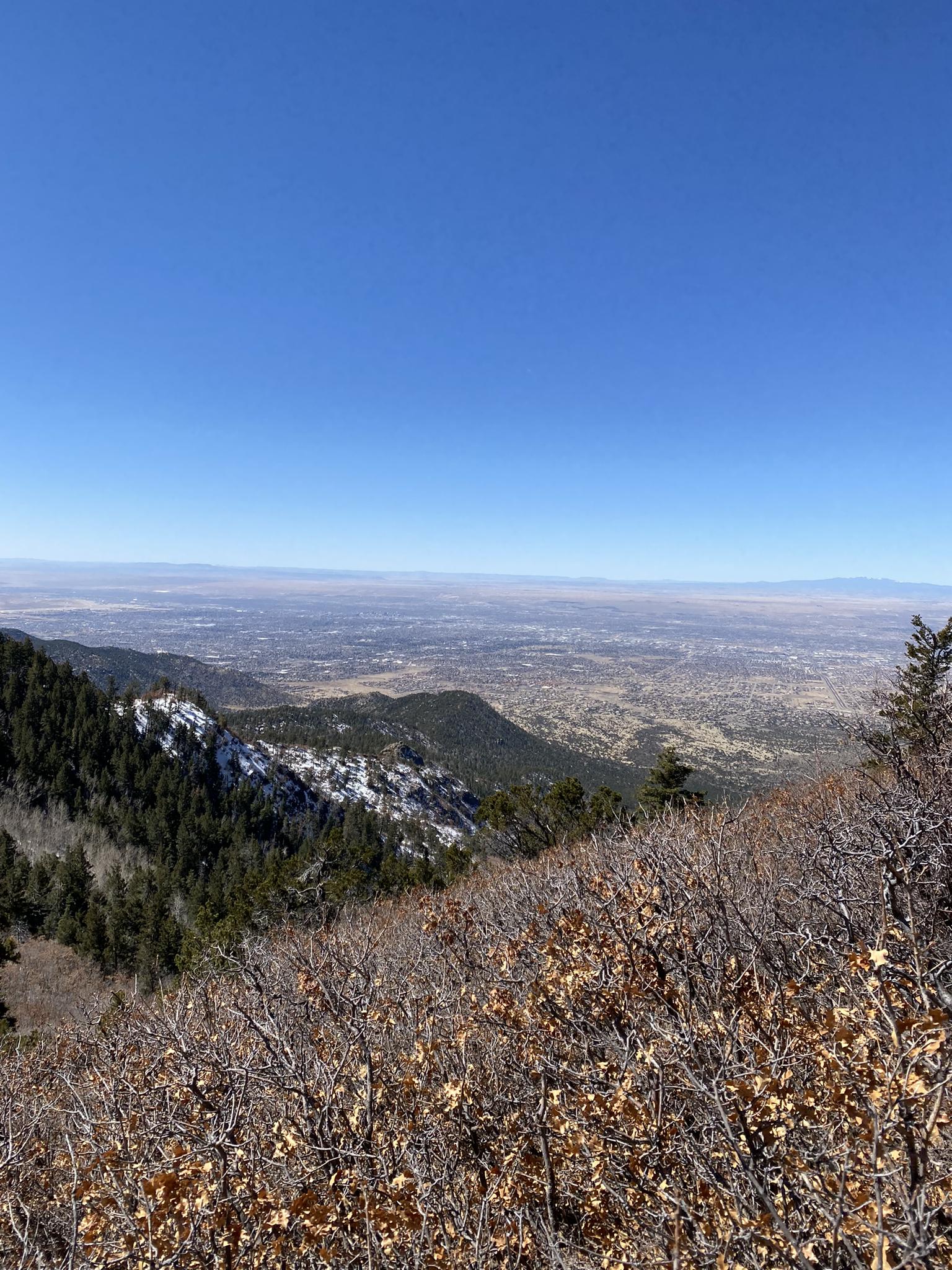 Mountain overlook with snow-dusted slopes, distant valley, and clear blue sky.