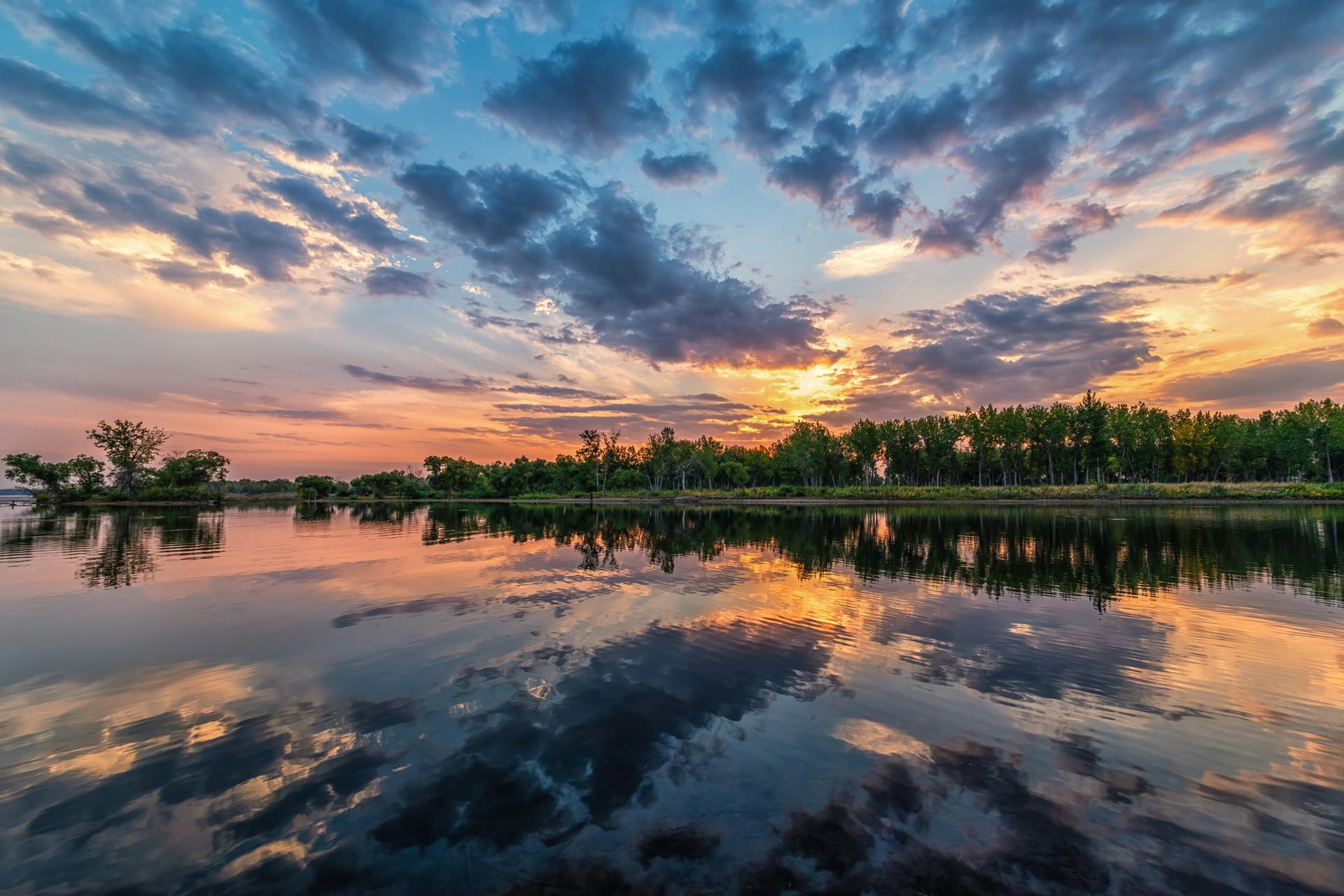 Vibrant sunset sky reflected in calm water with a distant treeline.