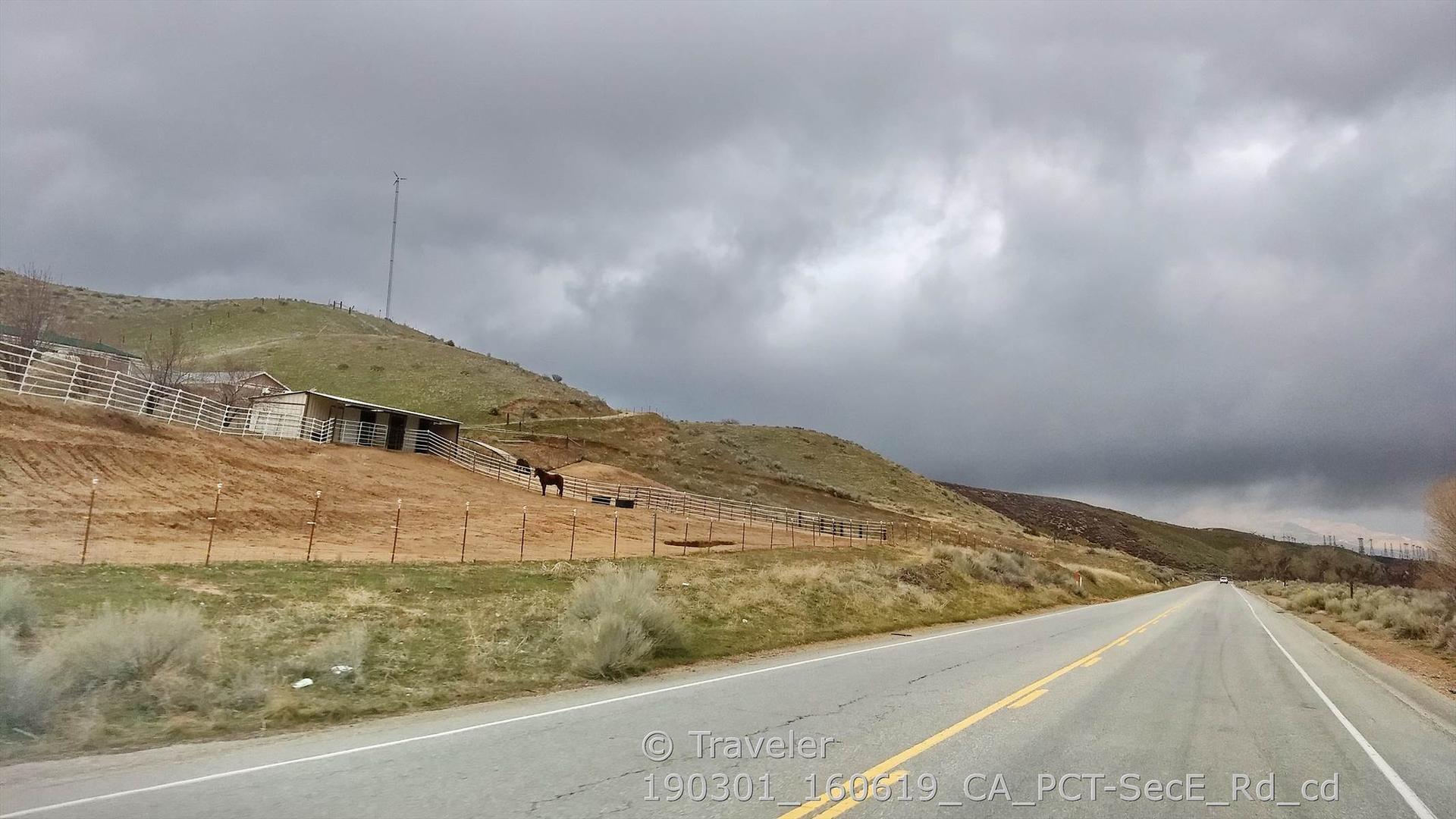 A road next to a fenced pasture with horses under a cloudy sky.