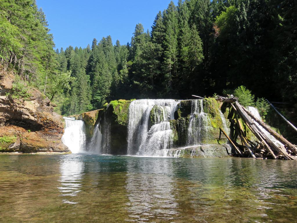 Wide waterfall flowing into a river, with lush green forest and blue sky.