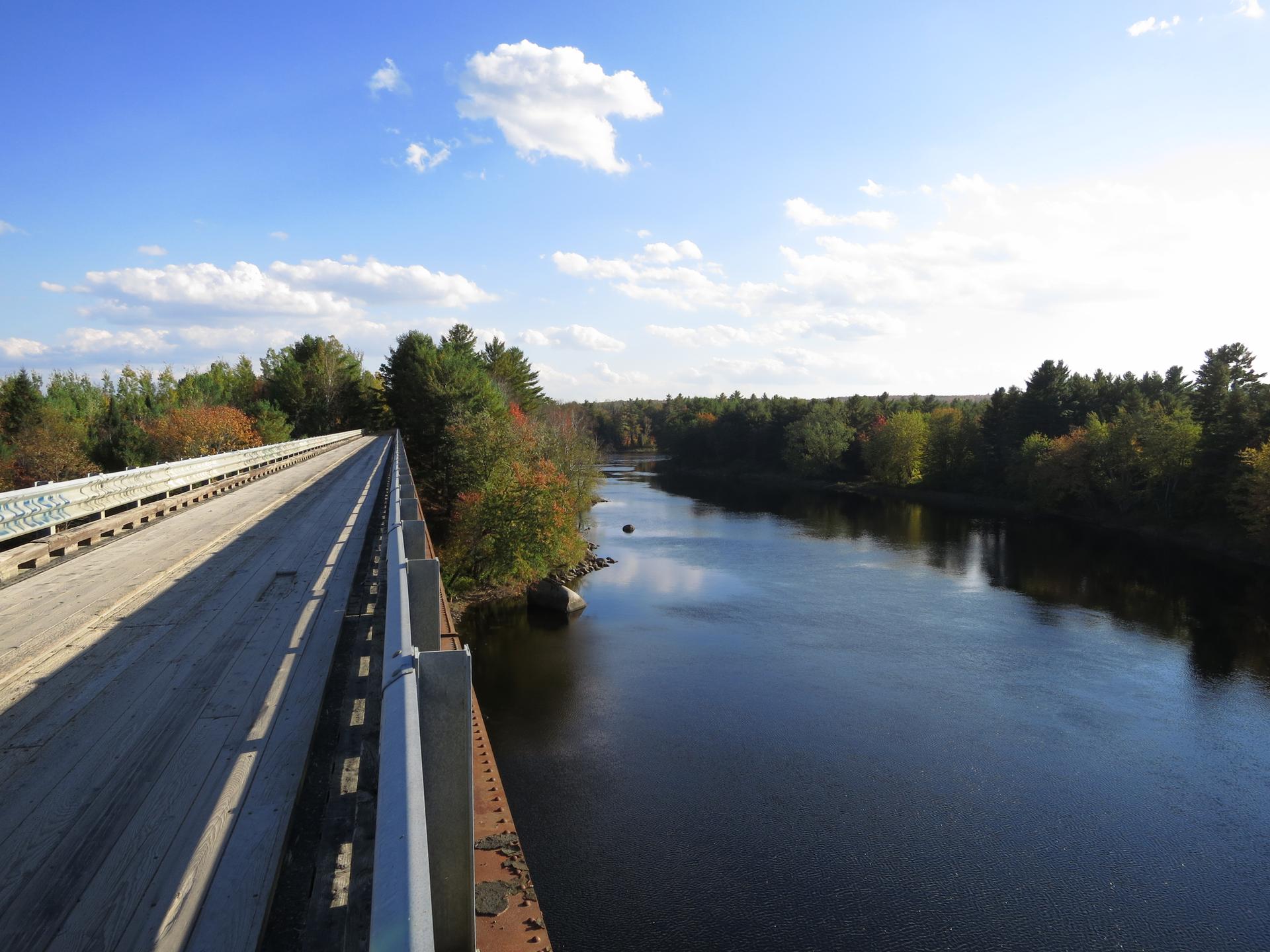 A long bridge spans a wide river, flanked by autumn trees under a sunny sky.