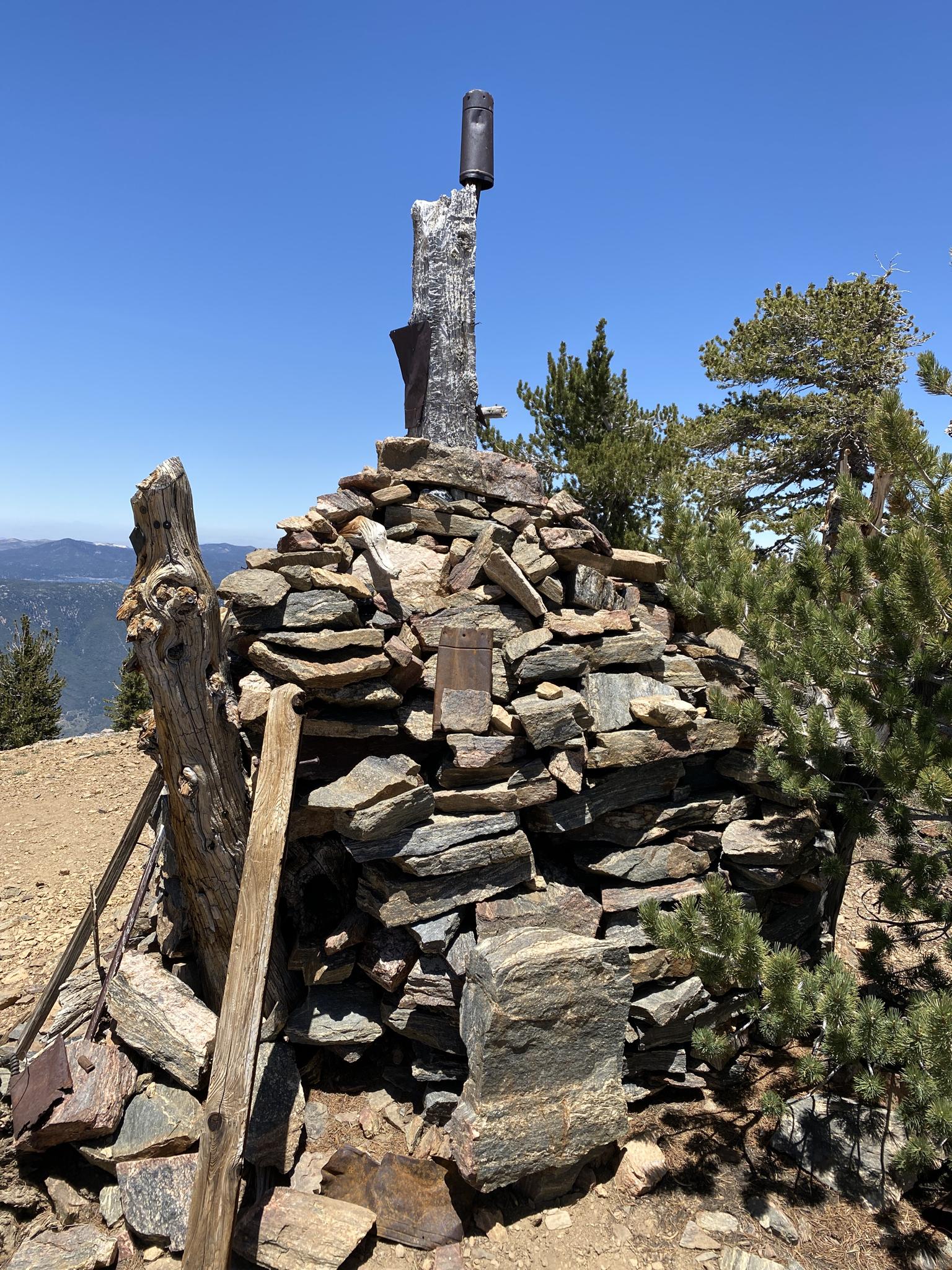 A rock cairn with a metal post on a mountain summit under bright sun.