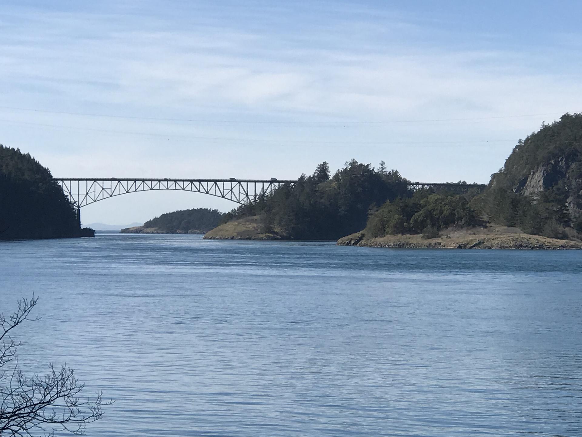 Sunny view of a wide water channel, forested islands, and a large steel bridge.