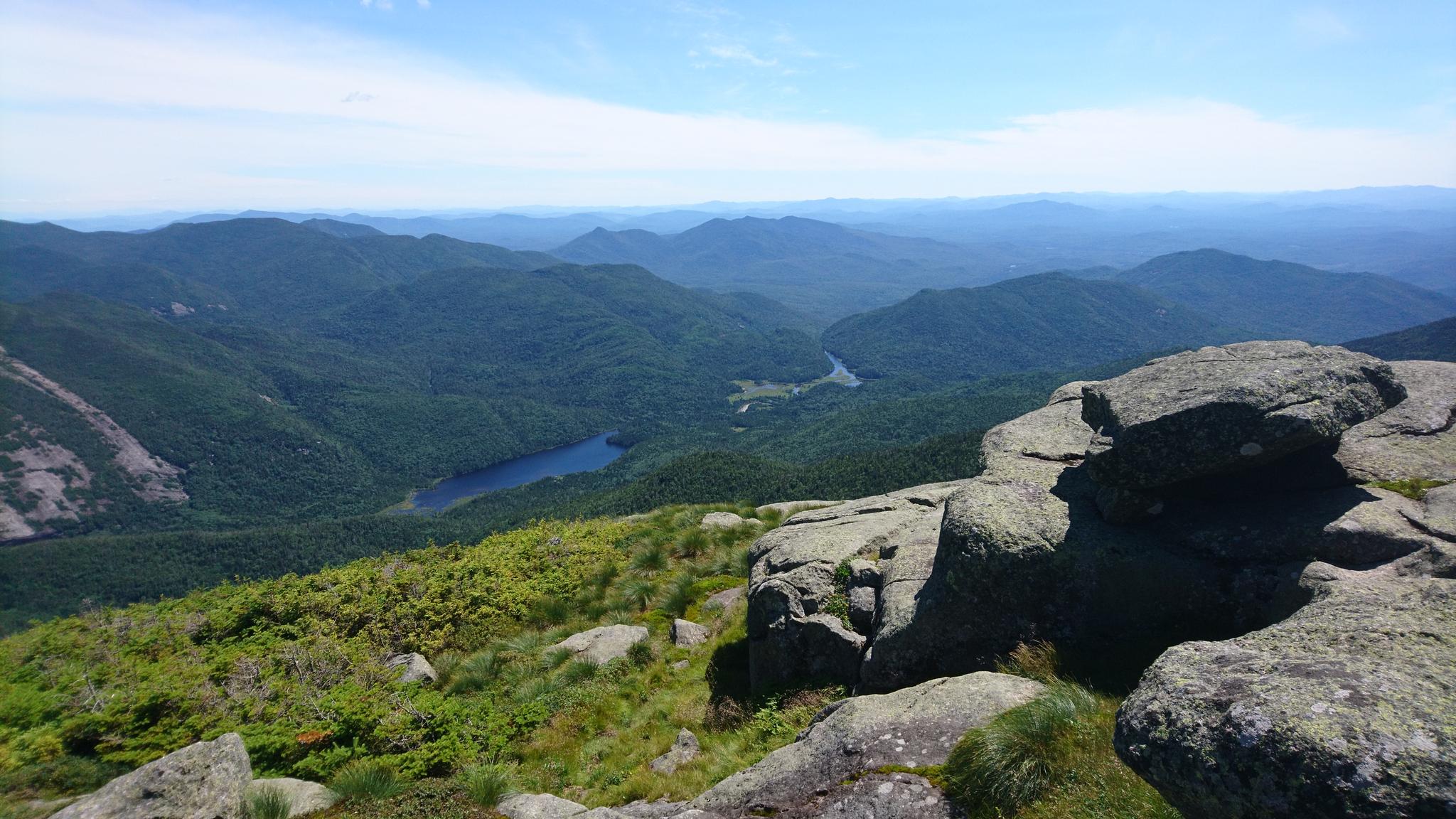 Panoramic mountain vista with a lake below, rocky foreground, under clear skies.