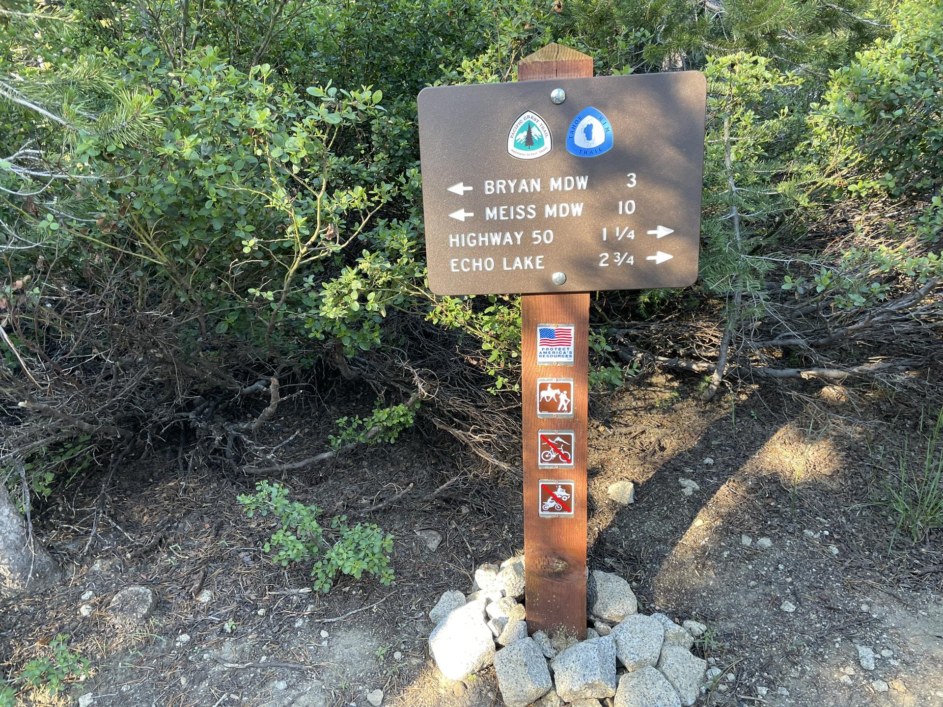Wooden trail sign with directional arrows and trail logos, surrounded by green bushes.