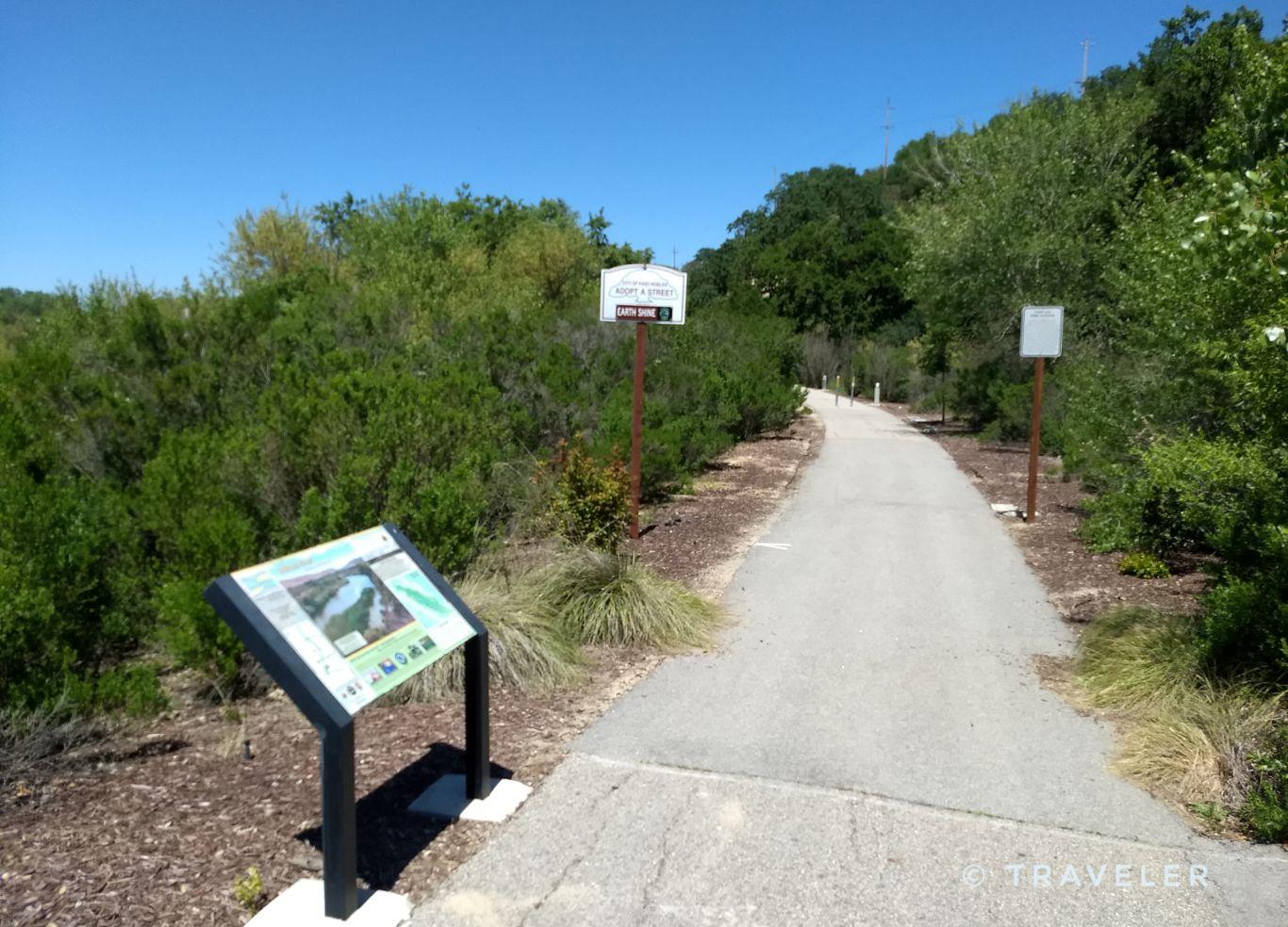A paved path with interpretive signs surrounded by dense green bushes on a sunny day.