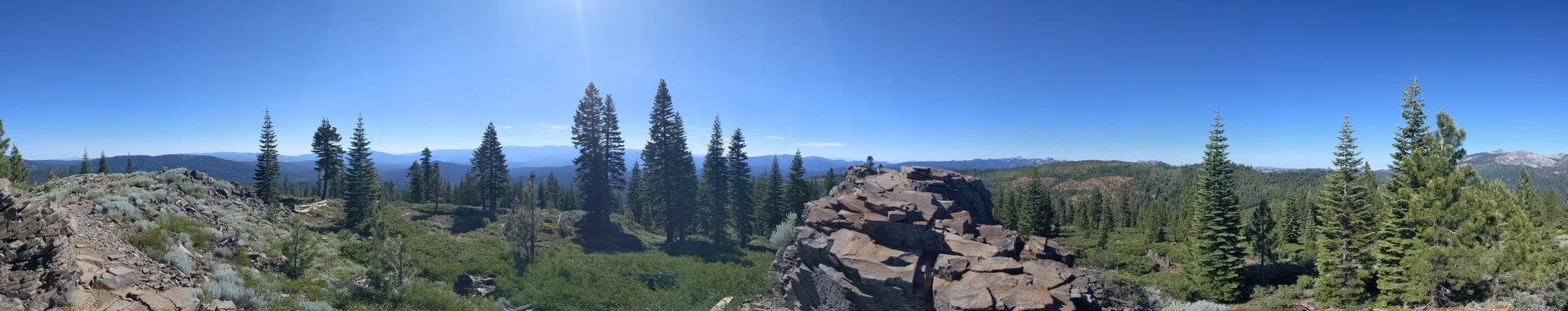 Panoramic view of a sunny mountain forest with a person on a rocky outcrop.