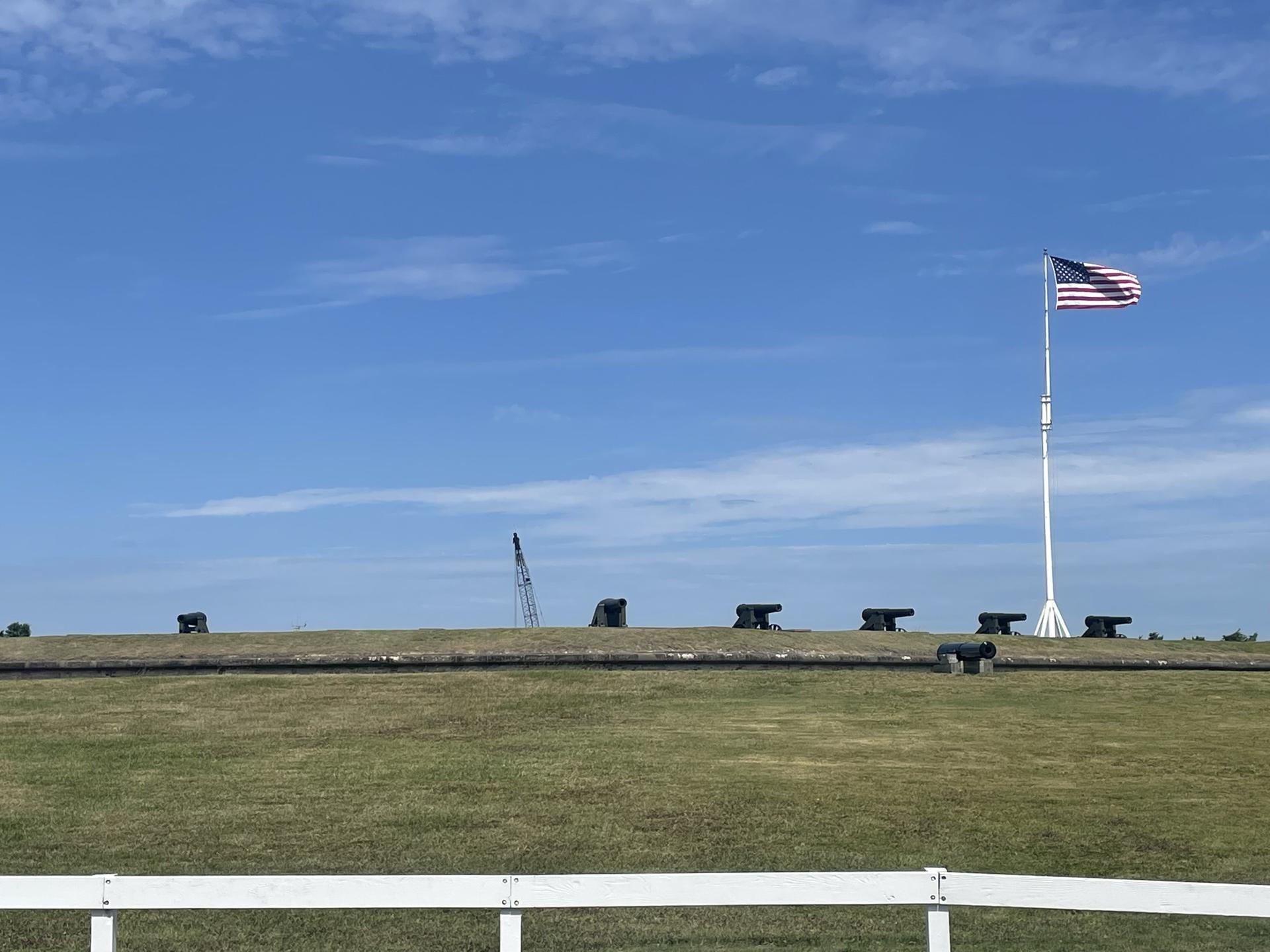 Cannons line a grassy field under a clear blue sky, with an American flag.