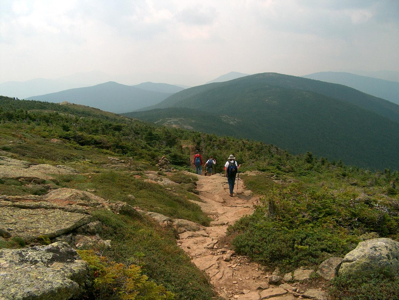 Three hikers ascend a rocky mountain trail under an overcast sky.
