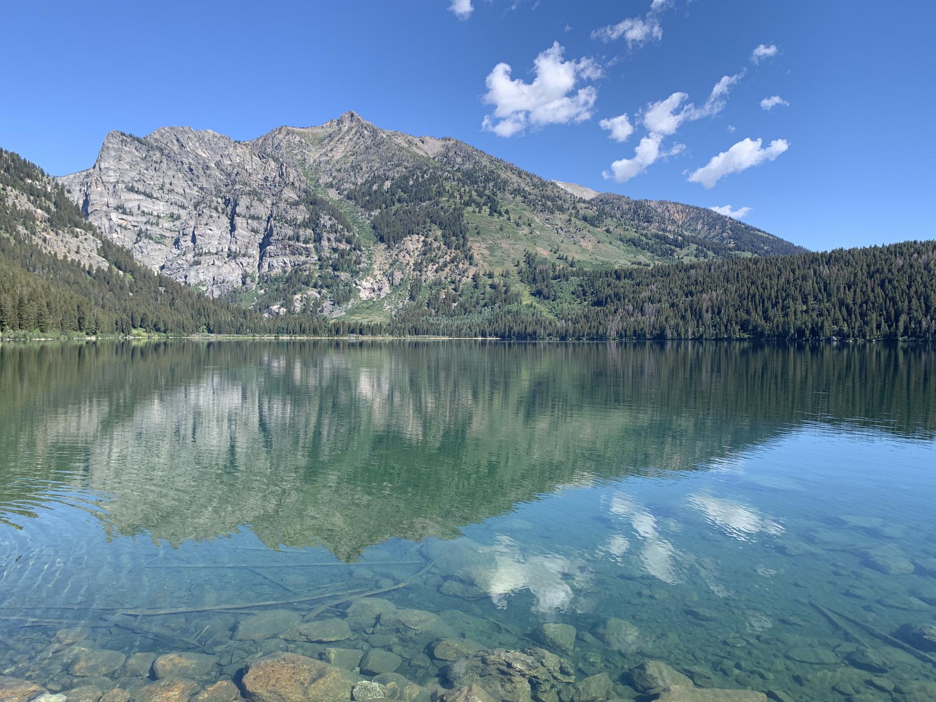 Clear lake reflecting mountains and sky, with visible rocks underwater.