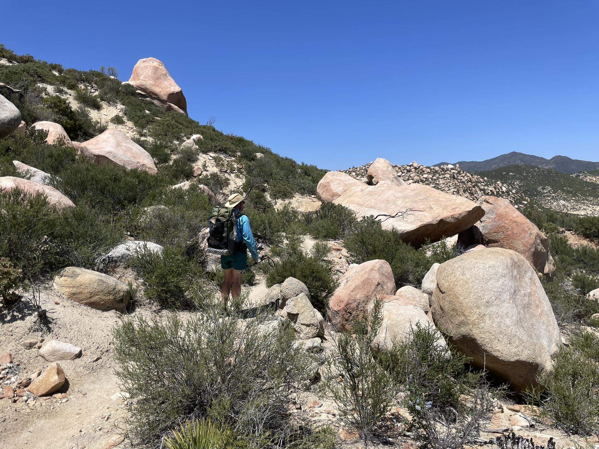 A hiker with a backpack walks a rocky trail on a sunny, boulder-strewn hillside.