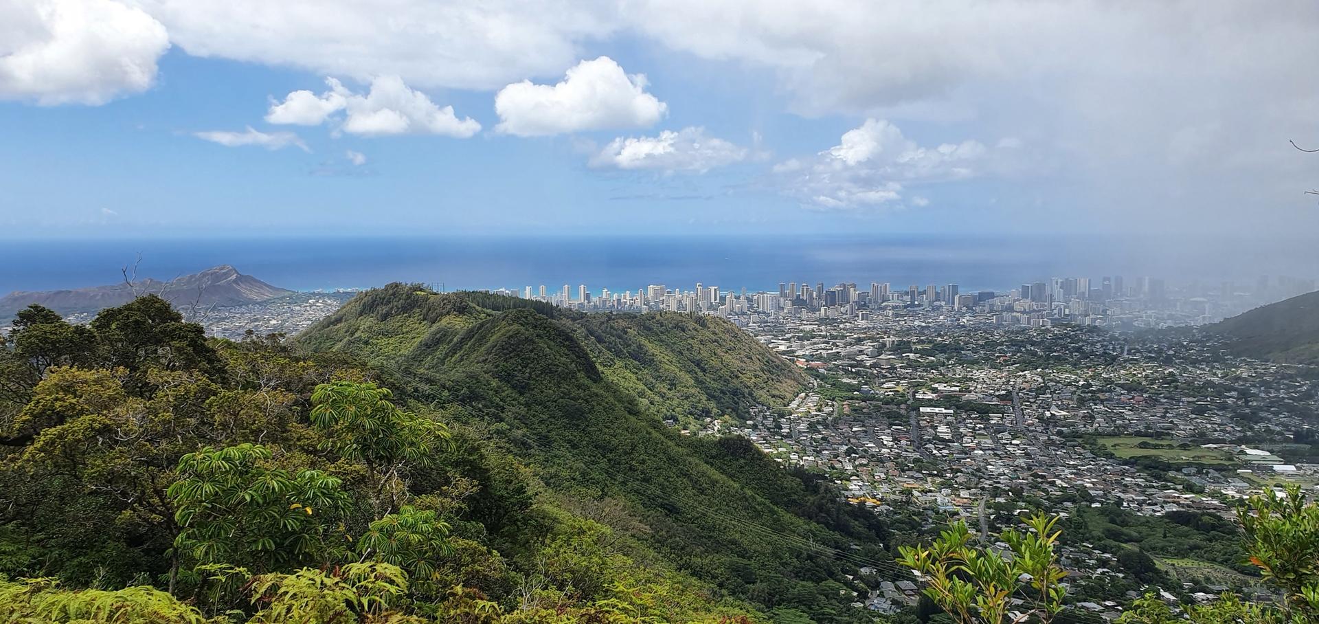 Panoramic view of a city, ocean, and green mountains under a bright, cloudy sky.