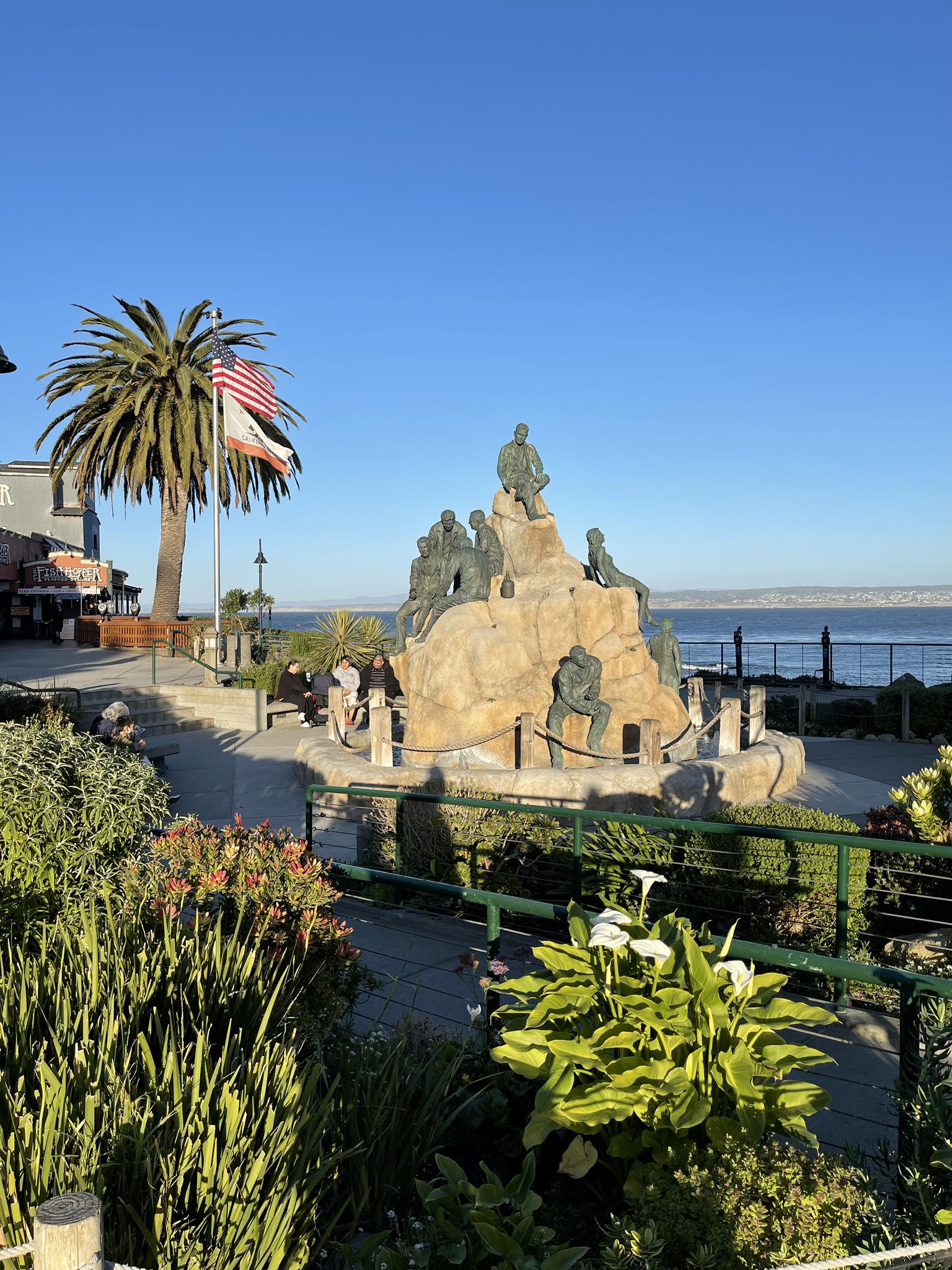 Bronze statue on a rock, with a palm tree, flags, and ocean view.