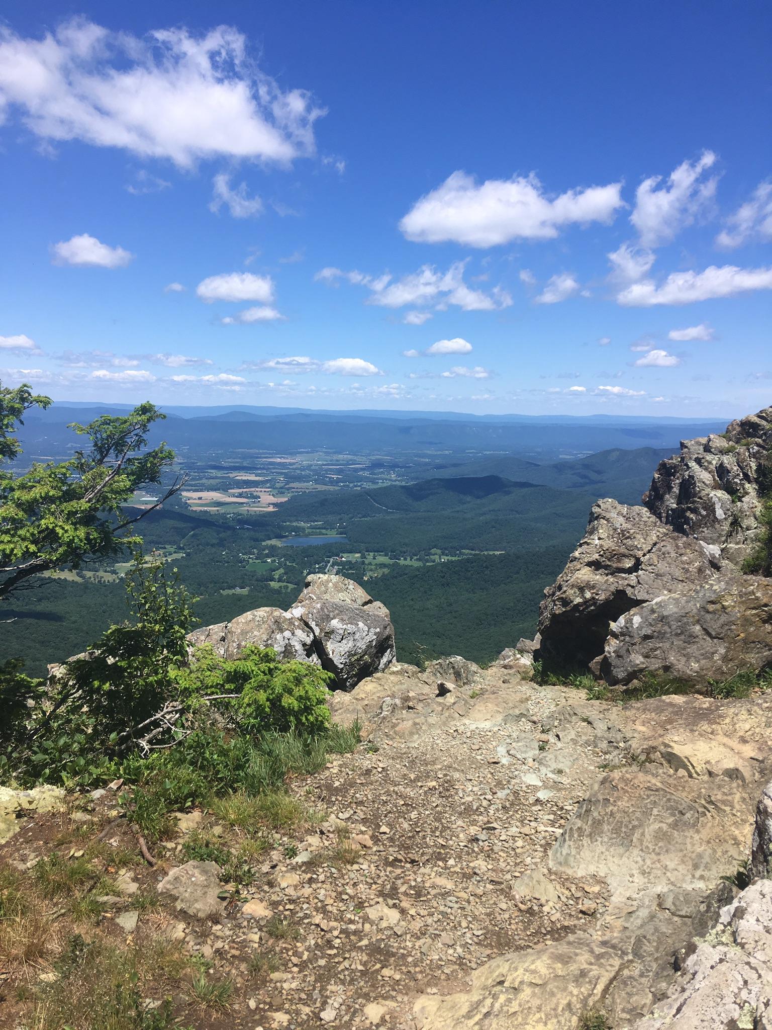 Rocky cliff edge overlooks a vast green valley under a blue sky.