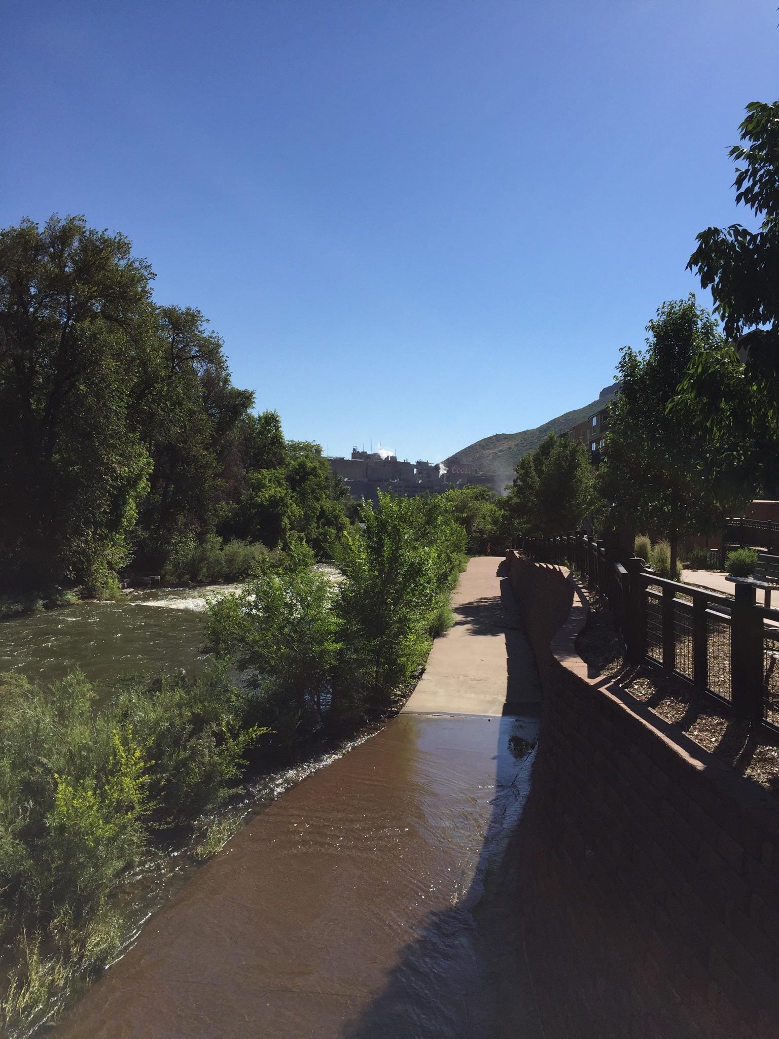 River flowing beside a paved path and wooden fence, surrounded by green trees.