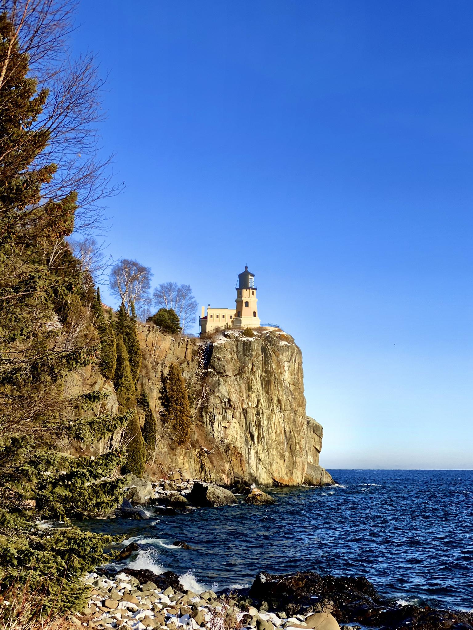 A white lighthouse stands atop a rocky cliff overlooking dark blue water.