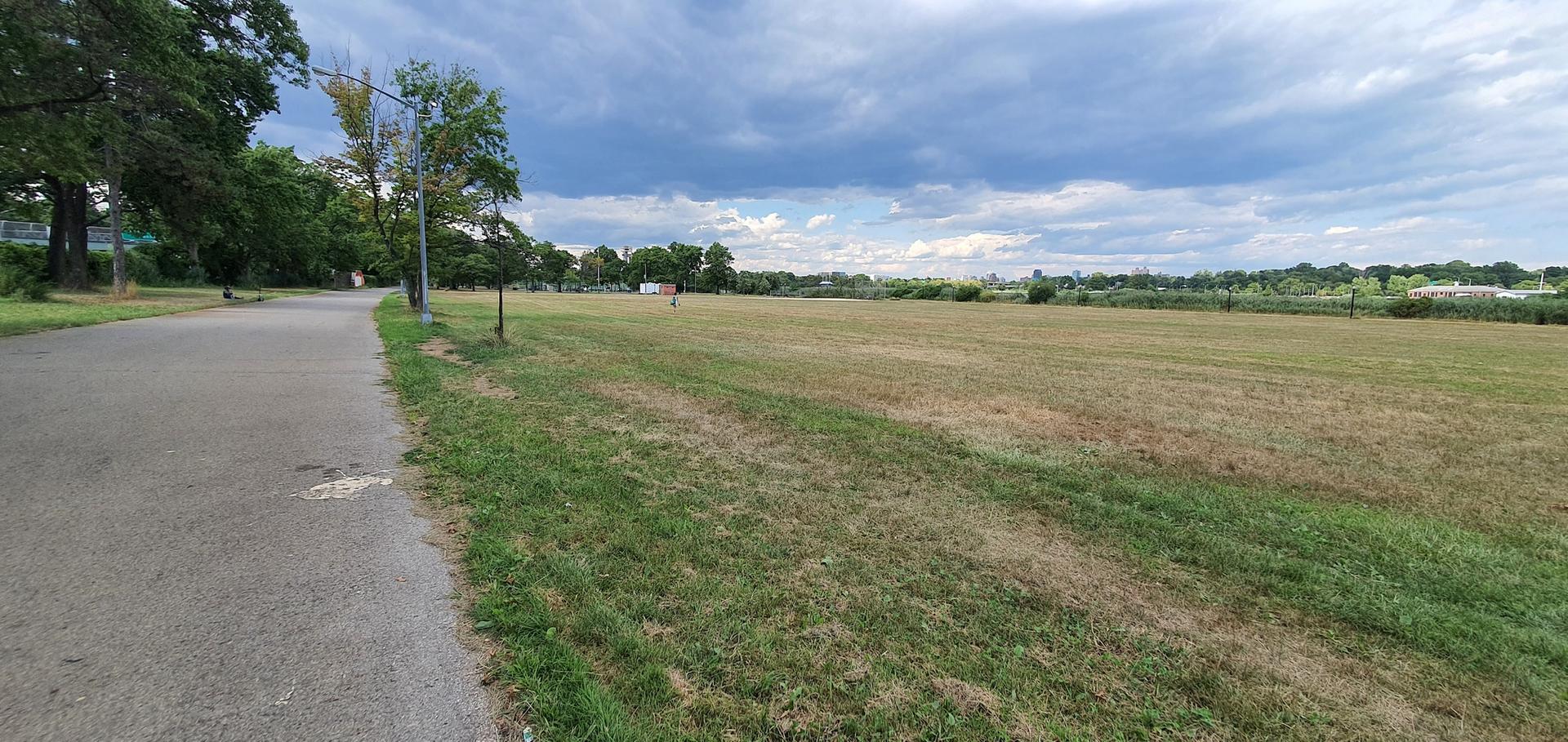 Wide open grassy field next to a paved path under a cloudy sky.