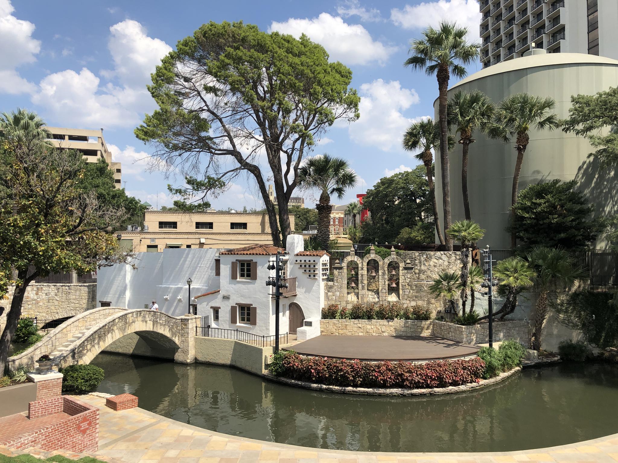 River with white building, arched bridge, outdoor stage, and palm trees.