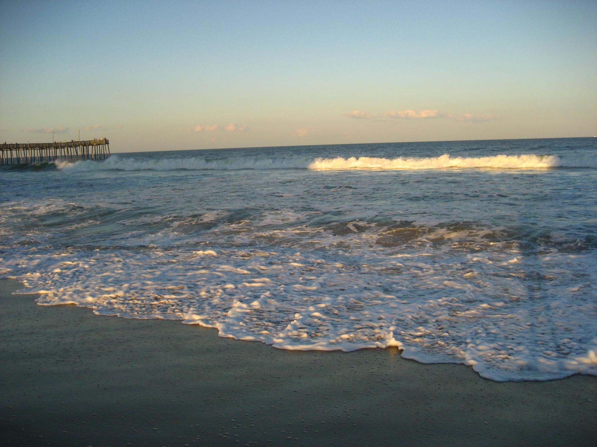 Ocean waves break on a sandy shore, a pier visible in the distance.