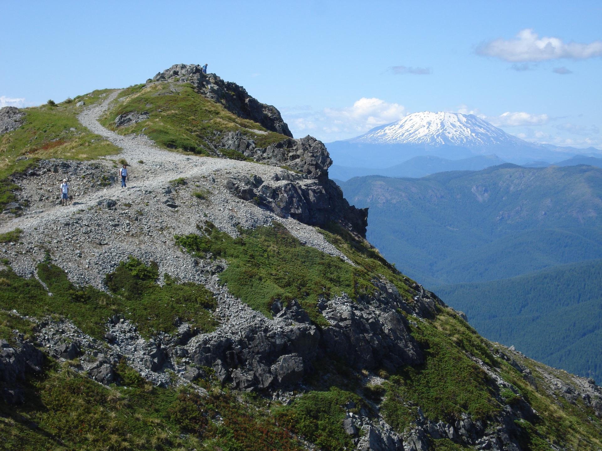 Two hikers on a rocky mountain trail with a distant snow-capped peak.