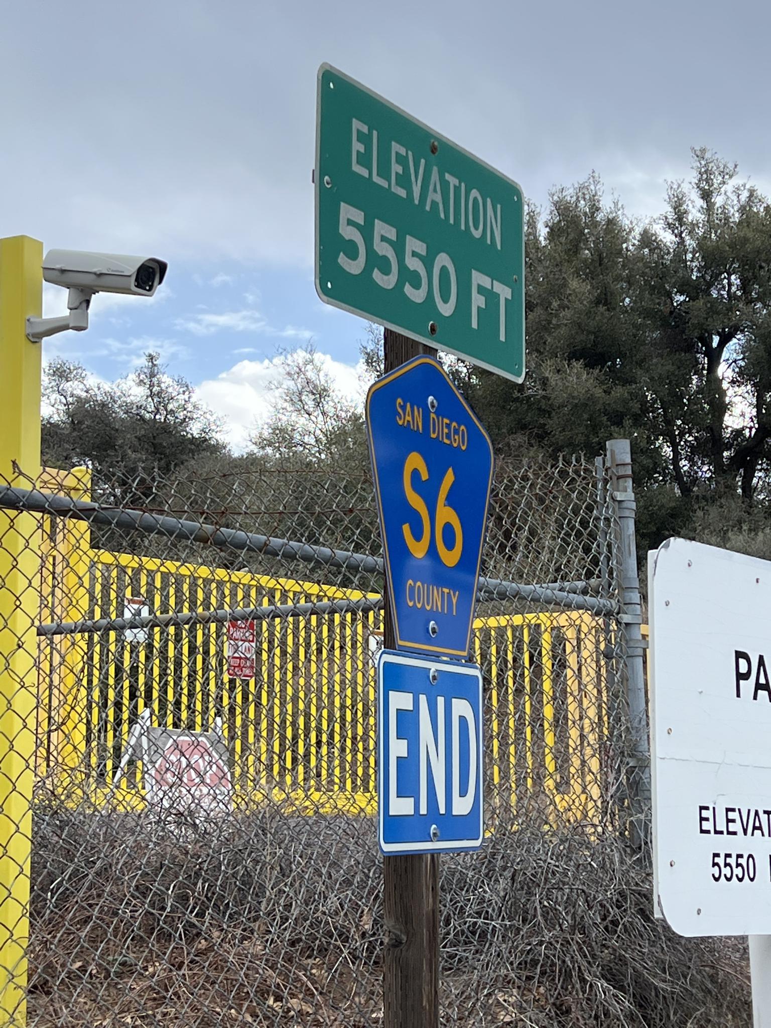 Road signs on a pole, including an elevation sign, next to a chain-link fence.