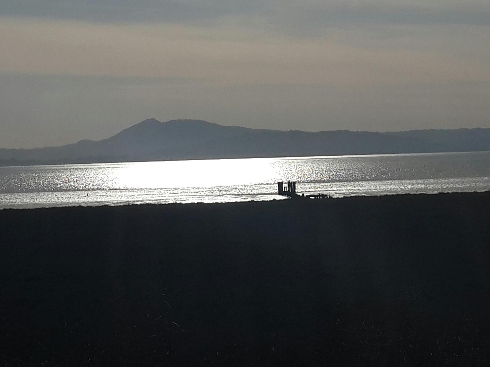 Sunlight reflects on water with distant mountains and a pier with people.