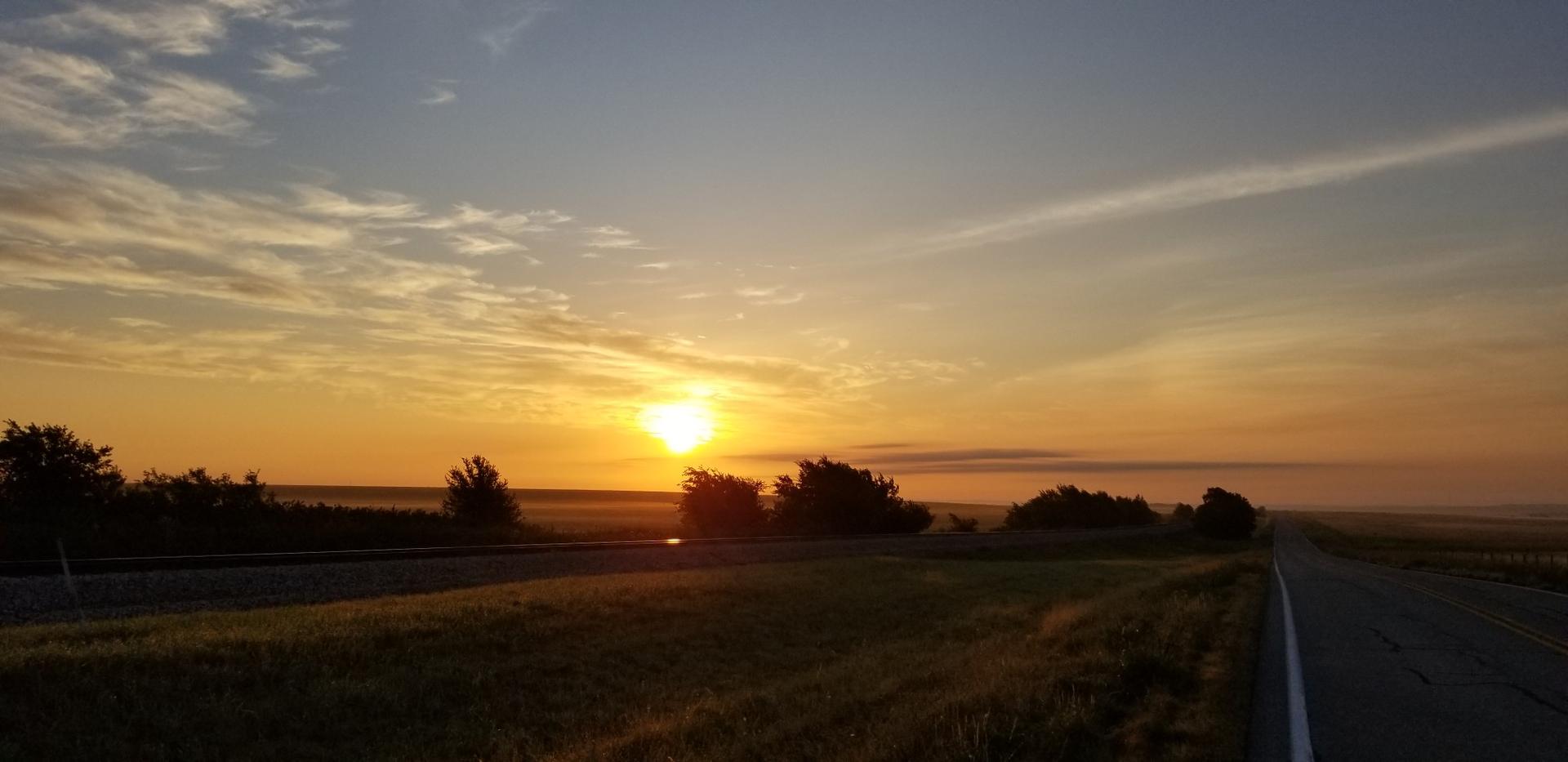 Golden sunrise over a rural road and field with distant trees.