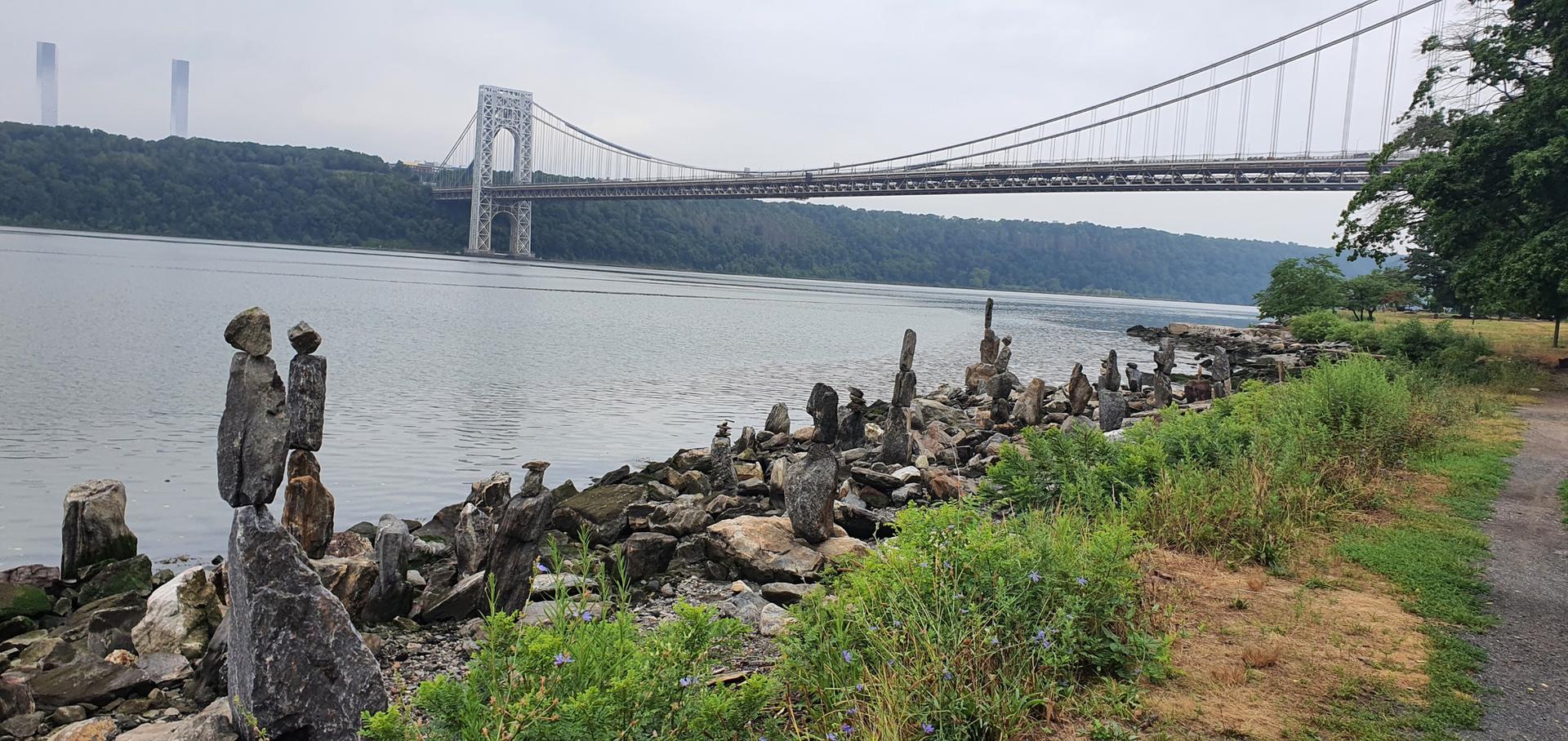 Stacked rock cairns line a riverbank with a large suspension bridge beyond.