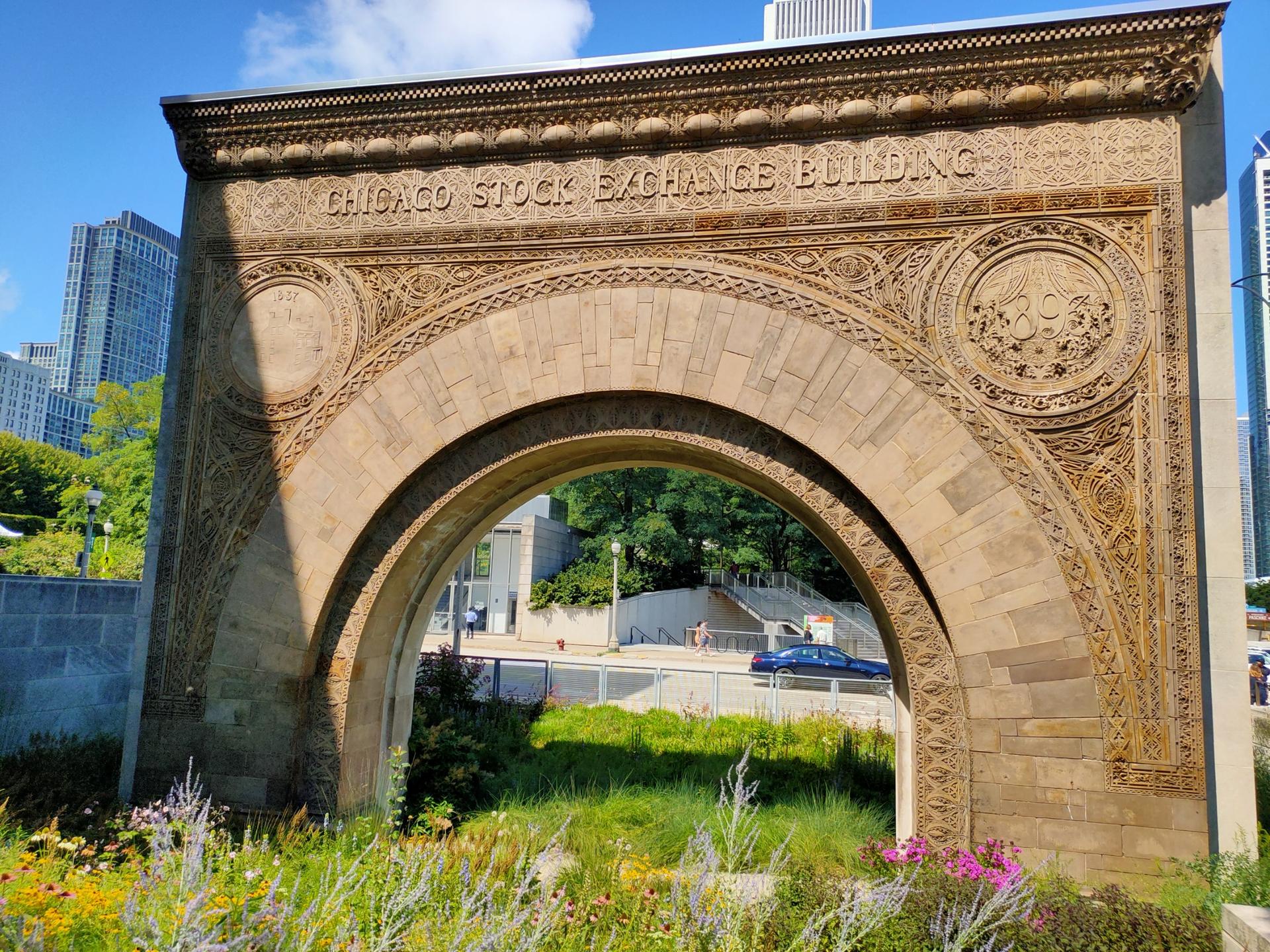 Ornate stone archway with text, surrounded by green plants under a sunny sky.