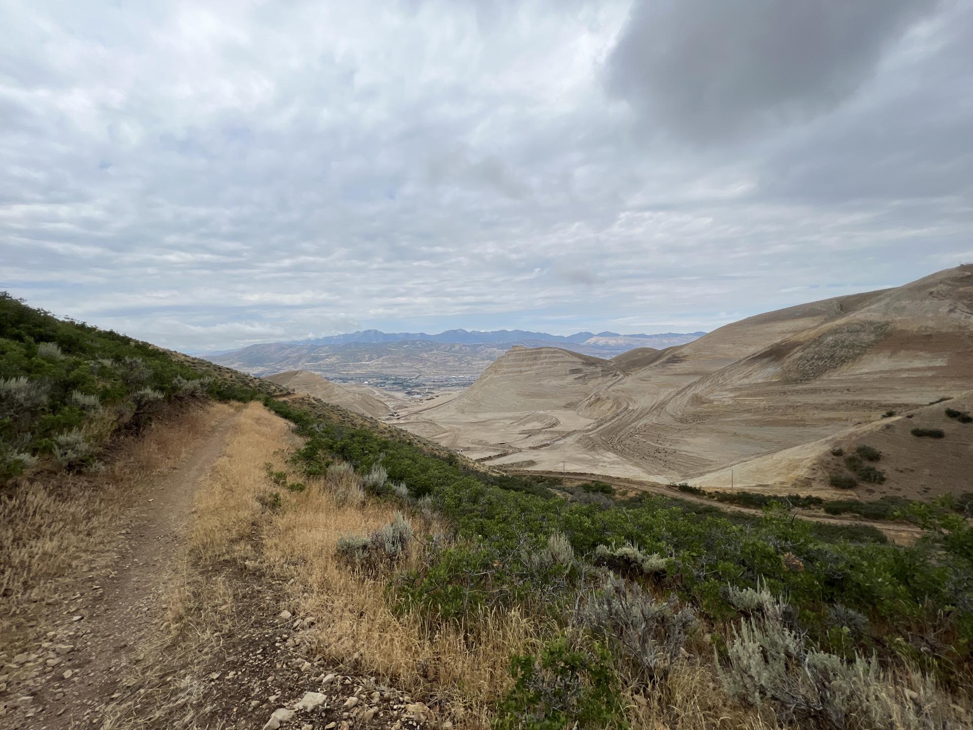 Dirt trail on a green hillside overlooking a dry valley under a cloudy sky.