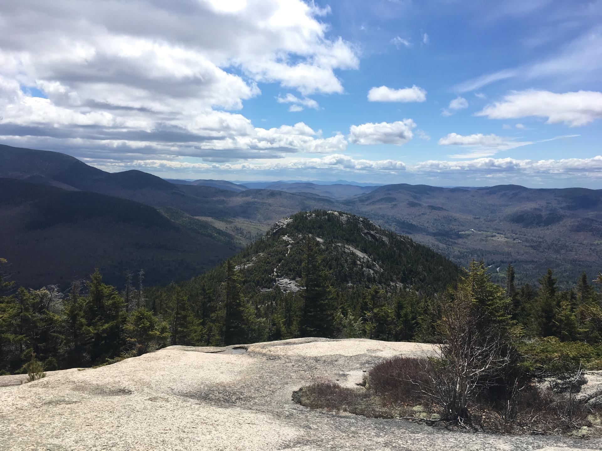 Expansive mountain vista with rocky foreground under a bright, cloudy sky.