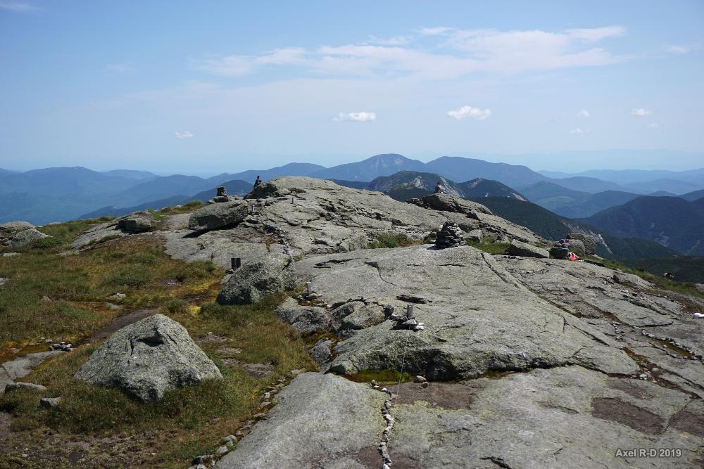 A rocky mountain summit with scattered cairns, overlooking distant blue peaks.