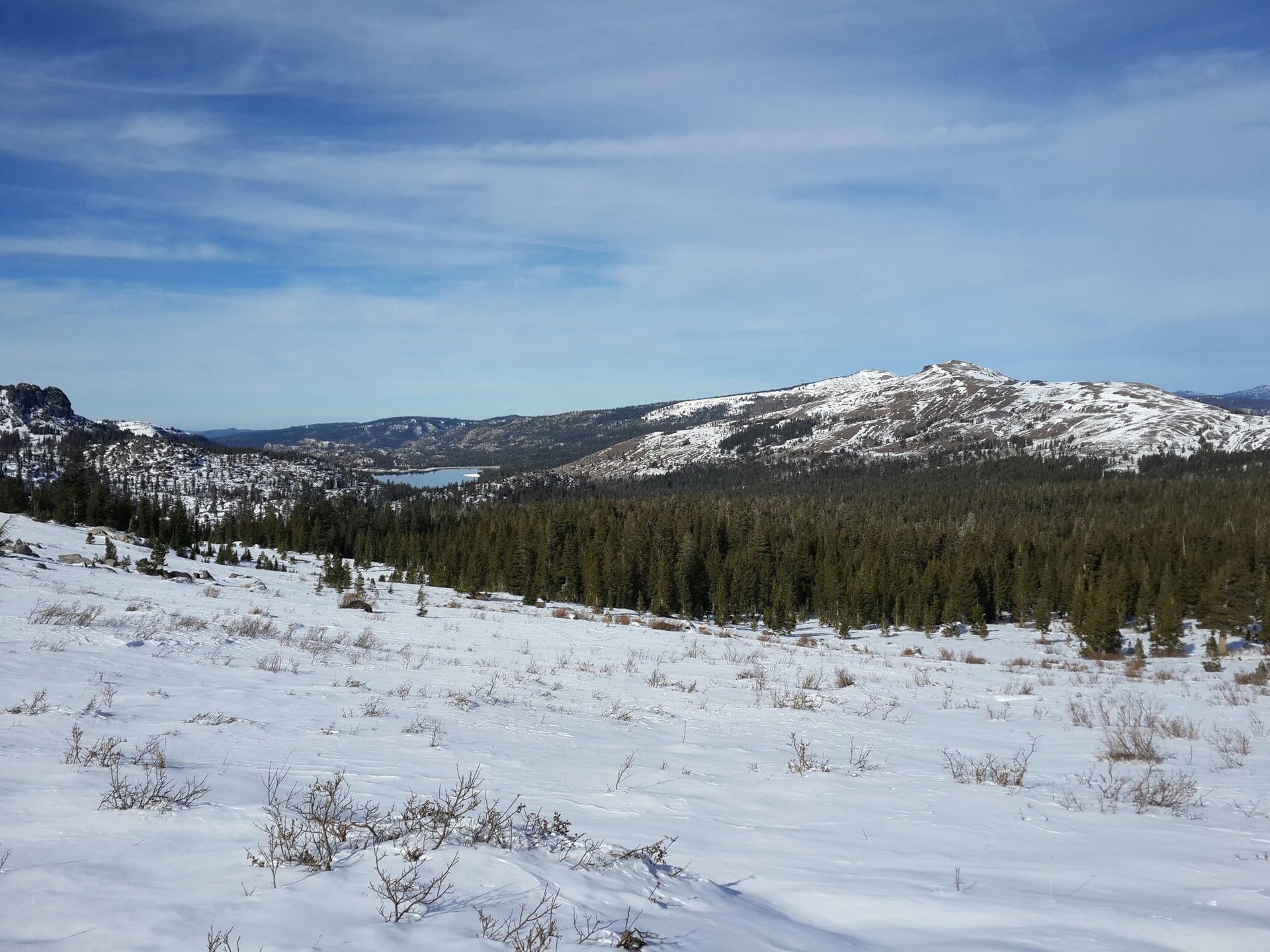 Expansive snow-covered landscape with distant lake, mountains, and blue sky.