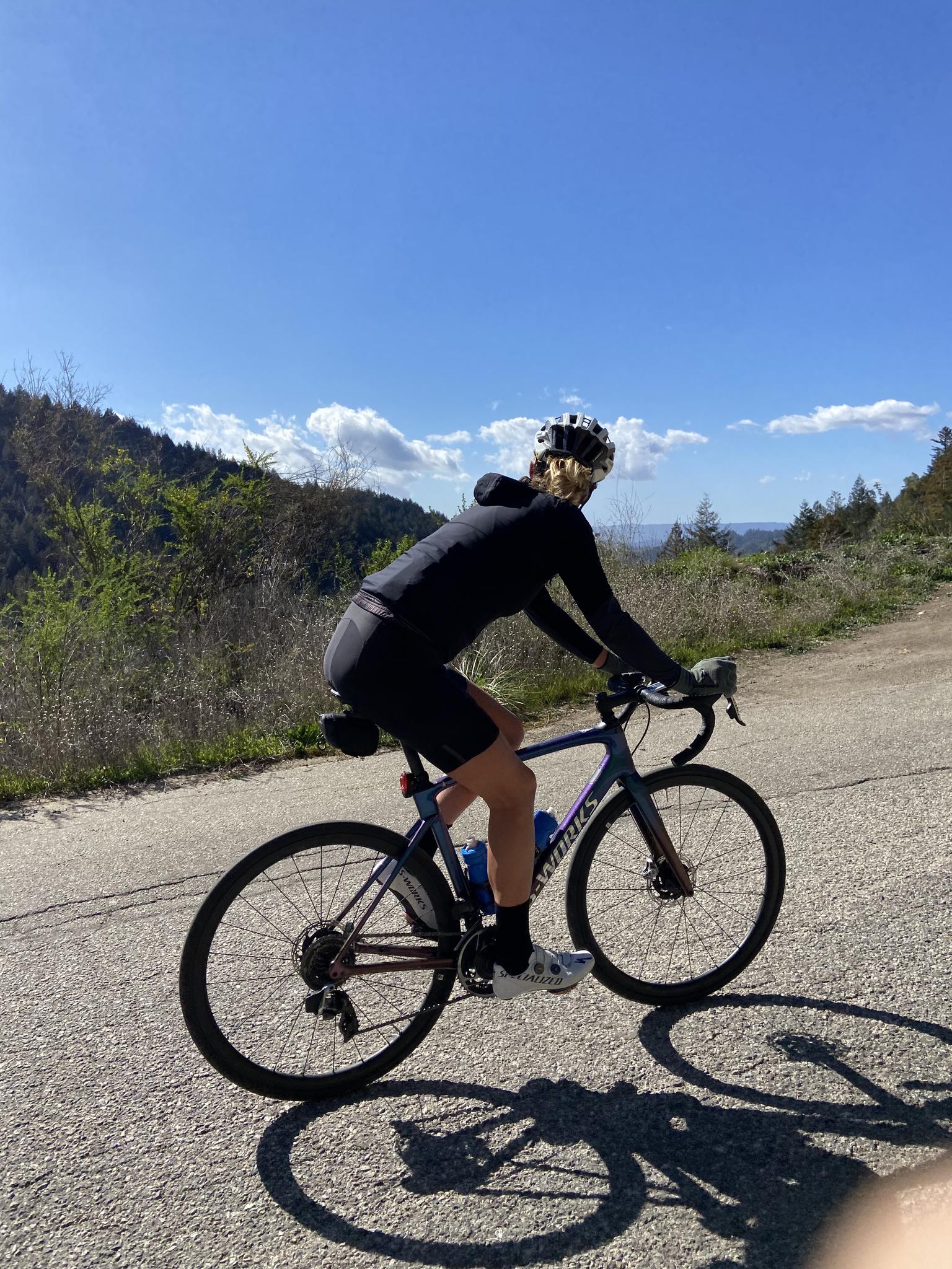 Woman cycling uphill on a sunny day, forested hills in the background.